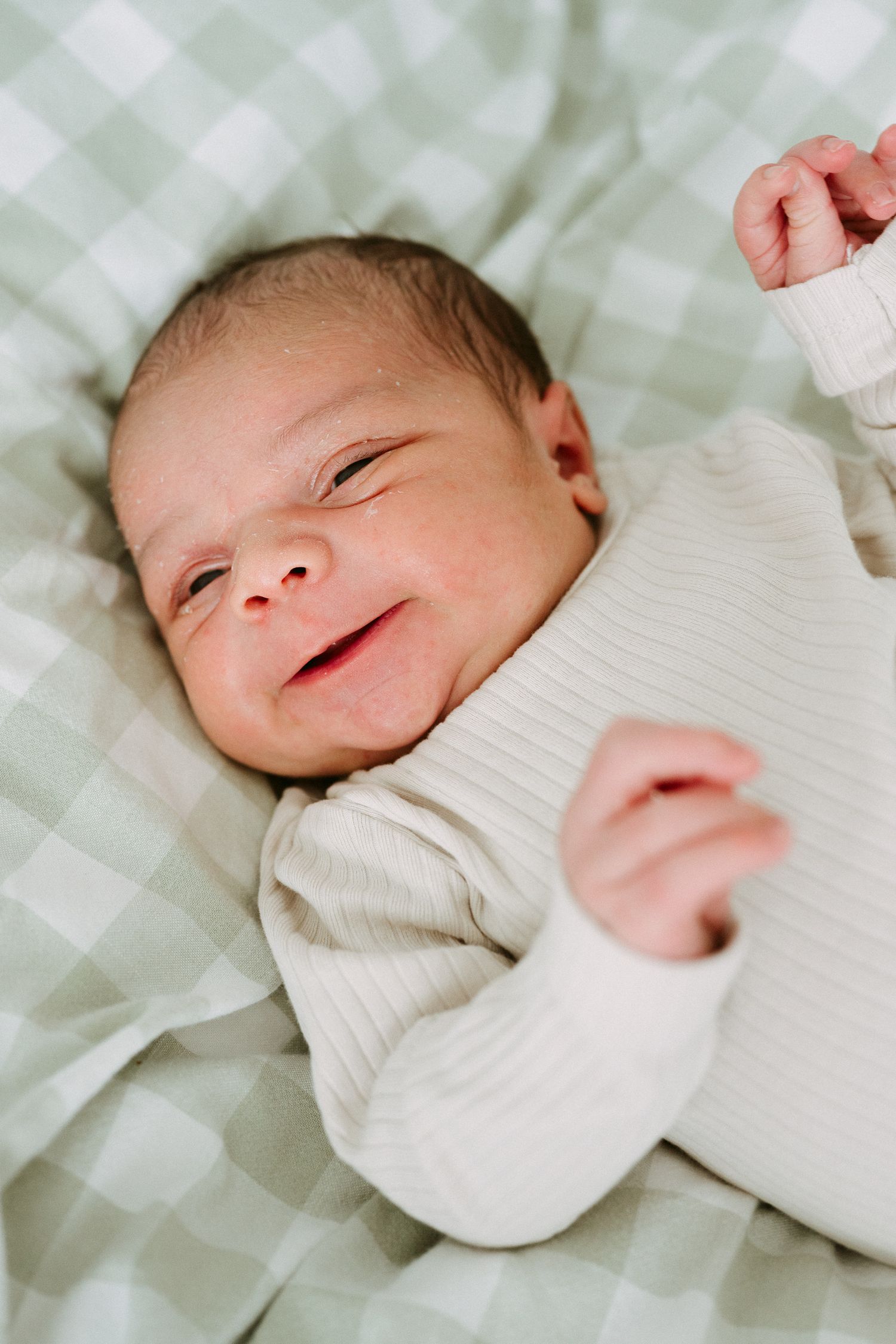 Smiling newborn baby in cream ribbed outfit lying on green gingham blanket.
