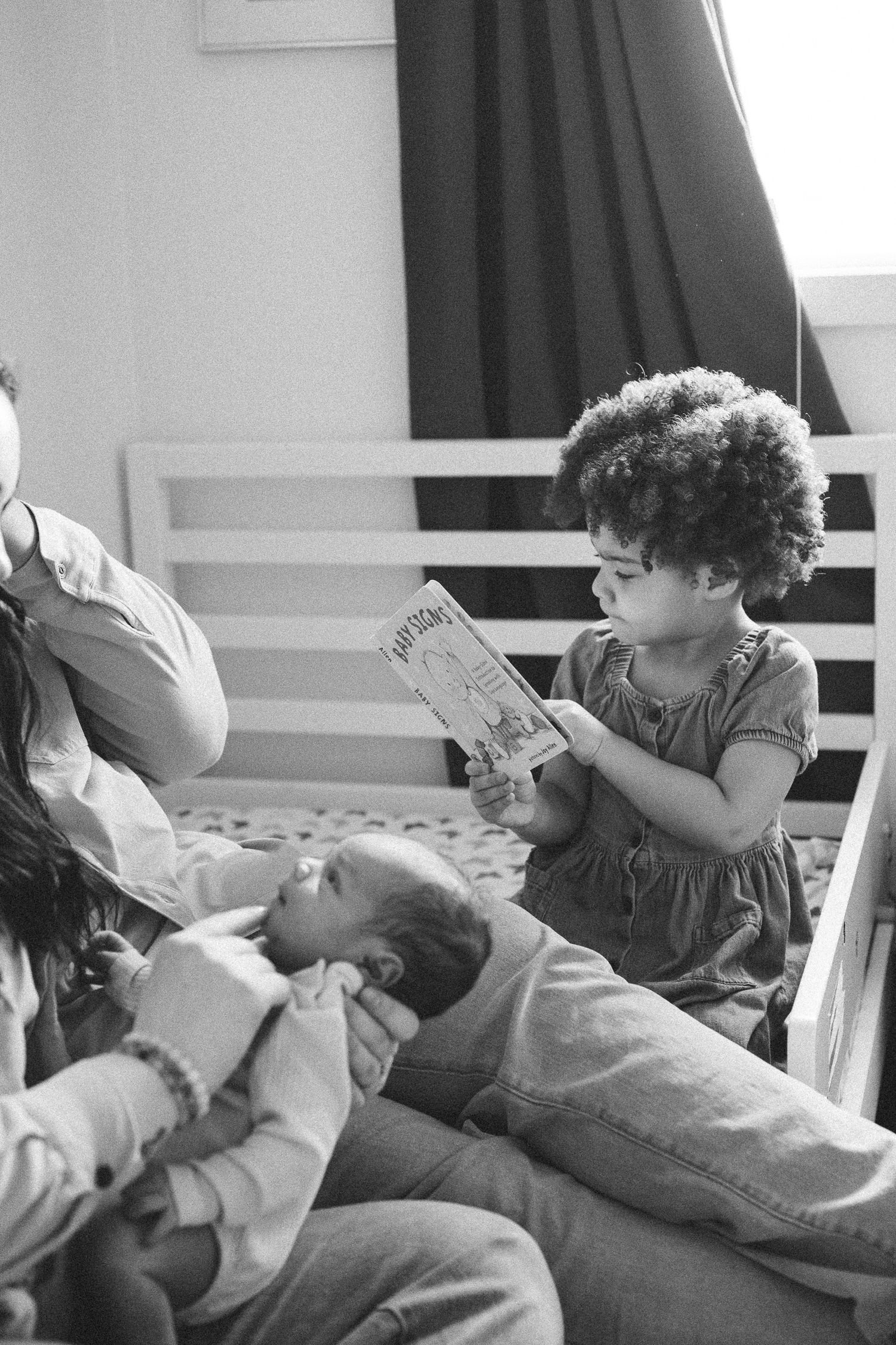 Black and white photo of curly-haired toddler reading a book to family members sitting on a bed.