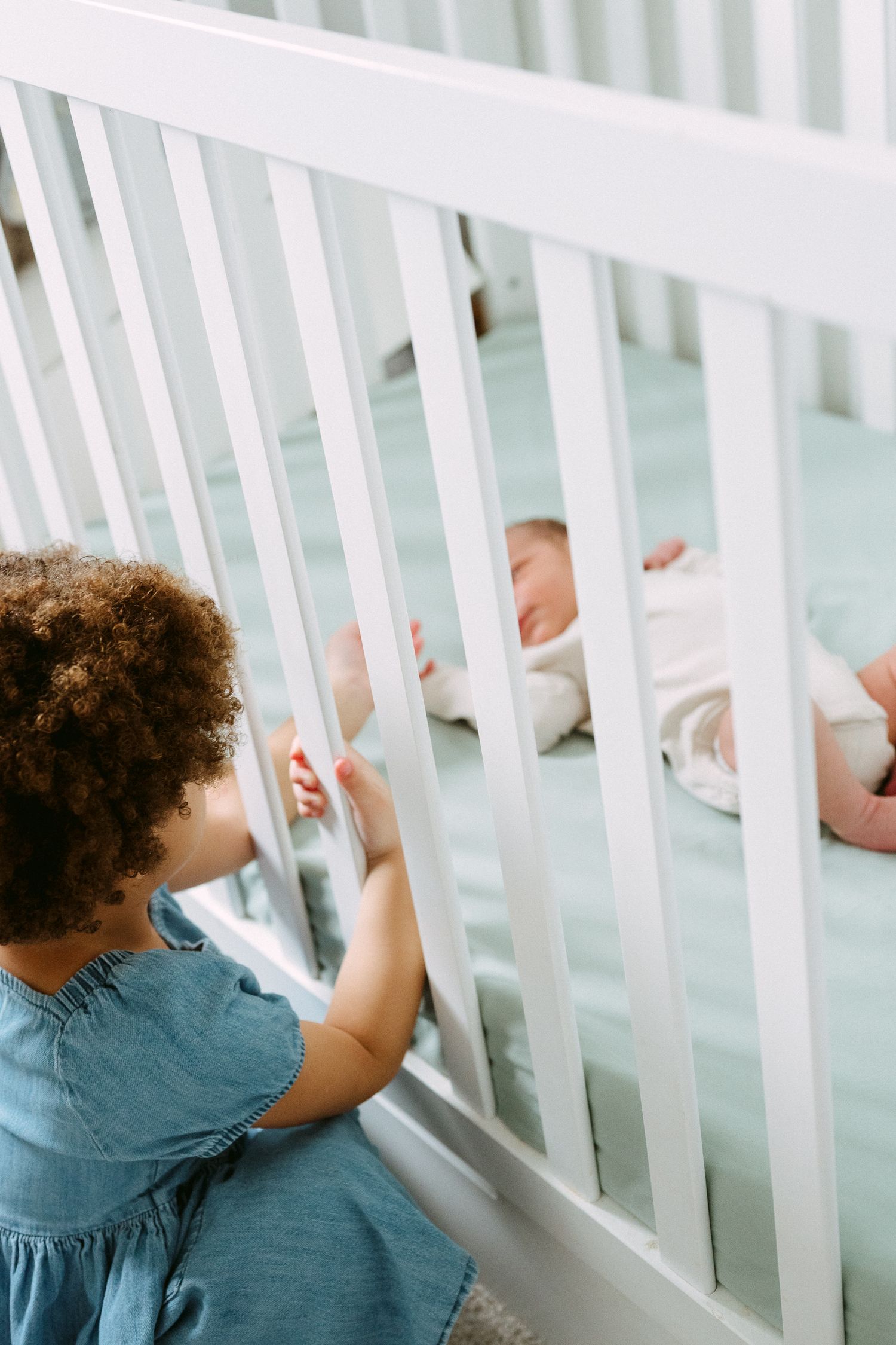 A young toddler in a blue dress looks through white crib rails at a resting newborn.