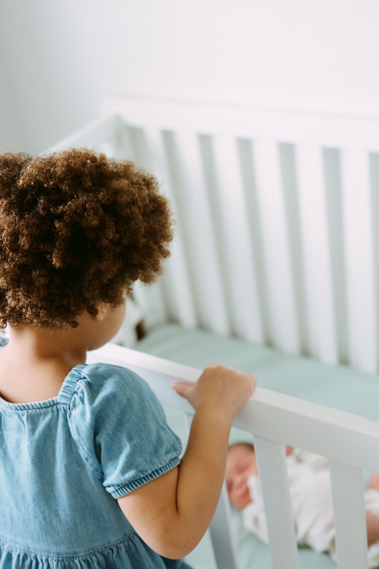 Young girl with curly hair in a blue dress looking over a white crib at a newborn baby.