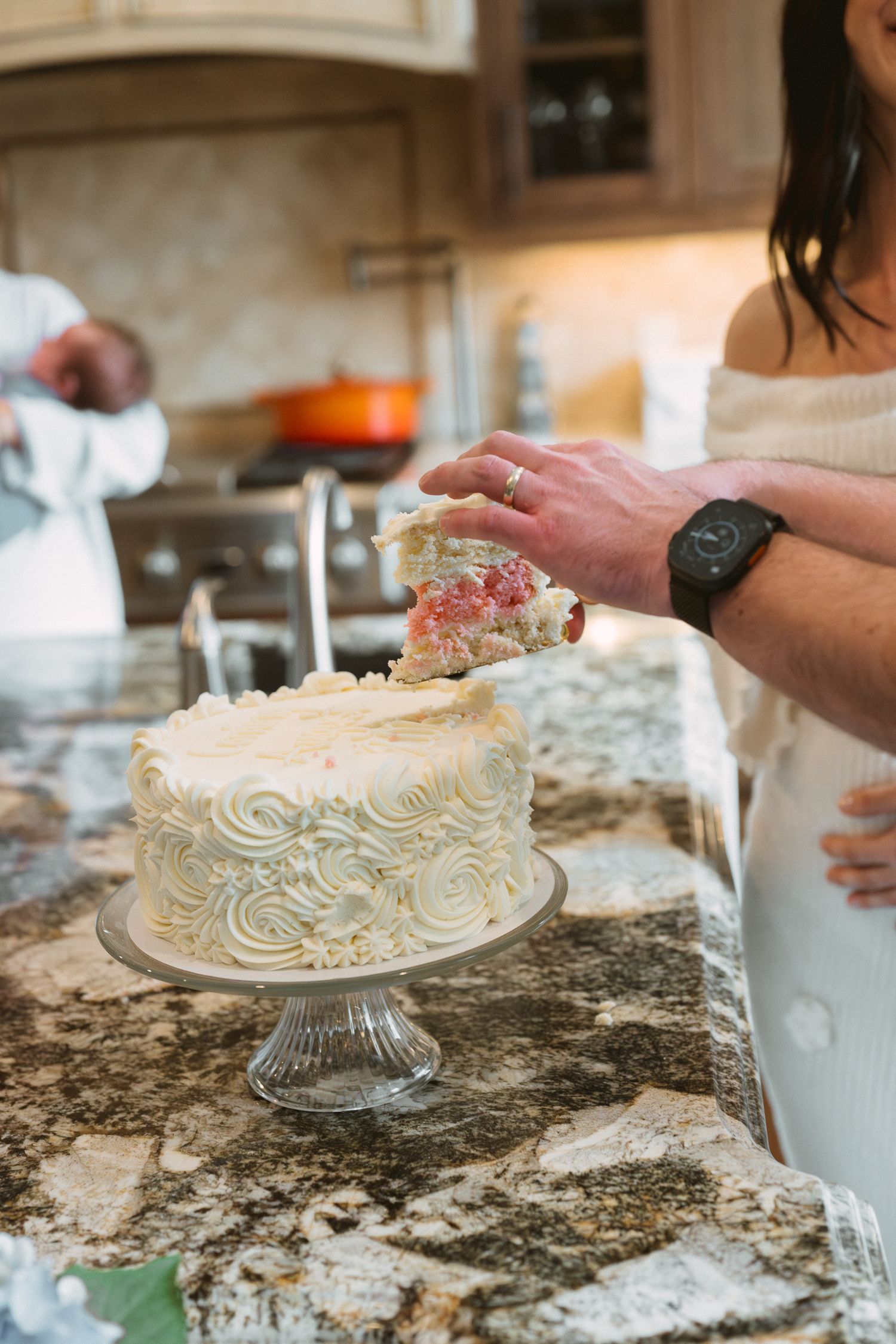 Couple cutting into white rosette frosted celebration cake on glass pedestal stand in kitchen.