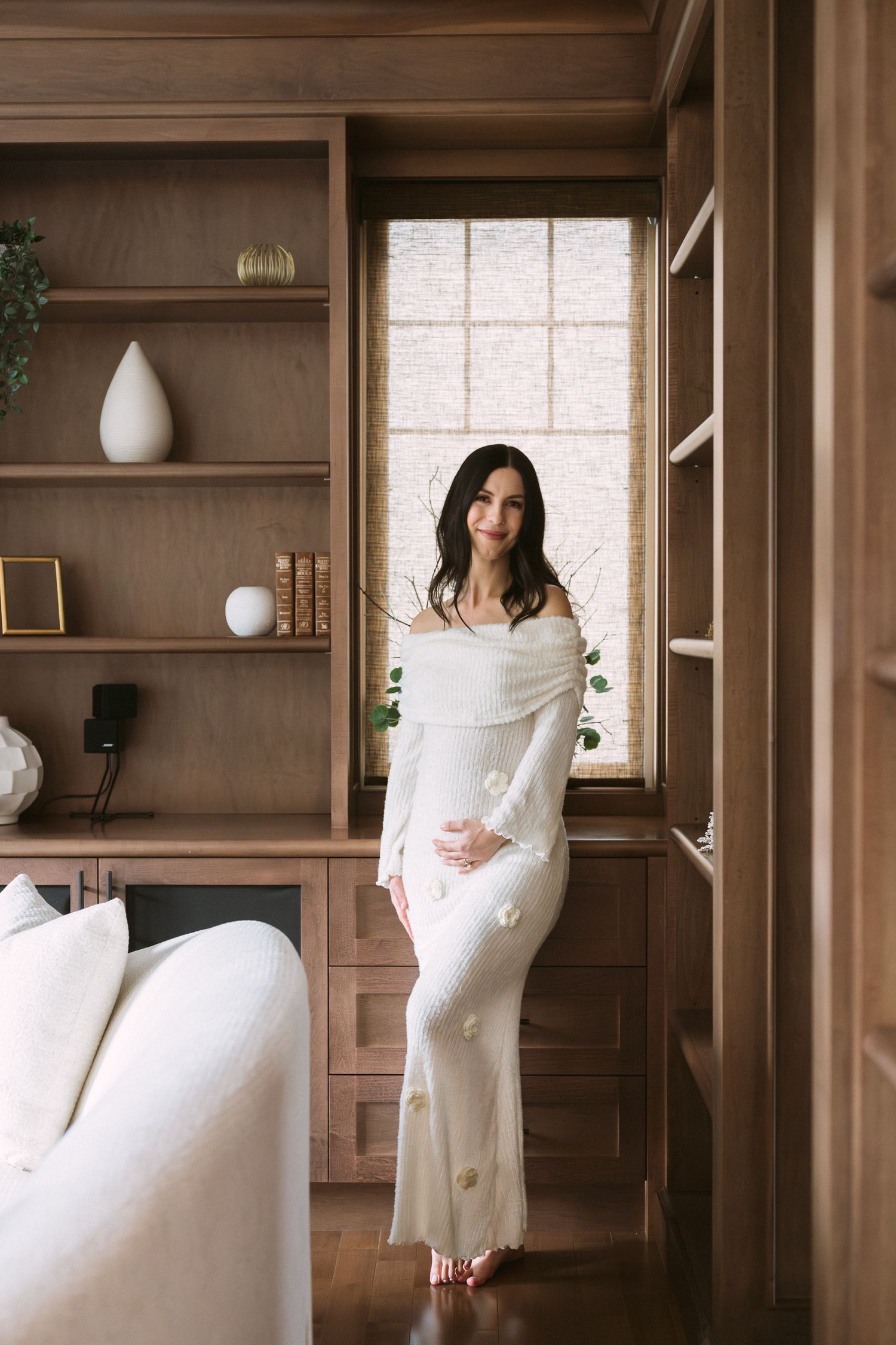 Pregnant woman in white off-shoulder floral maxi dress standing in elegant wood-paneled room with built-in shelves.