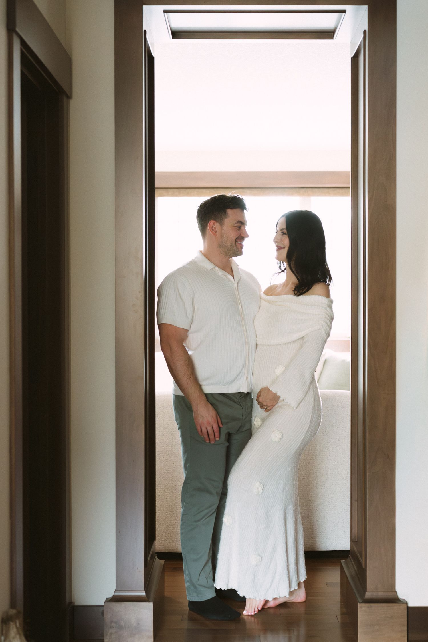 Couple sharing a loving glance in sunlit doorway, pregnant woman in elegant white knit dress barefoot.