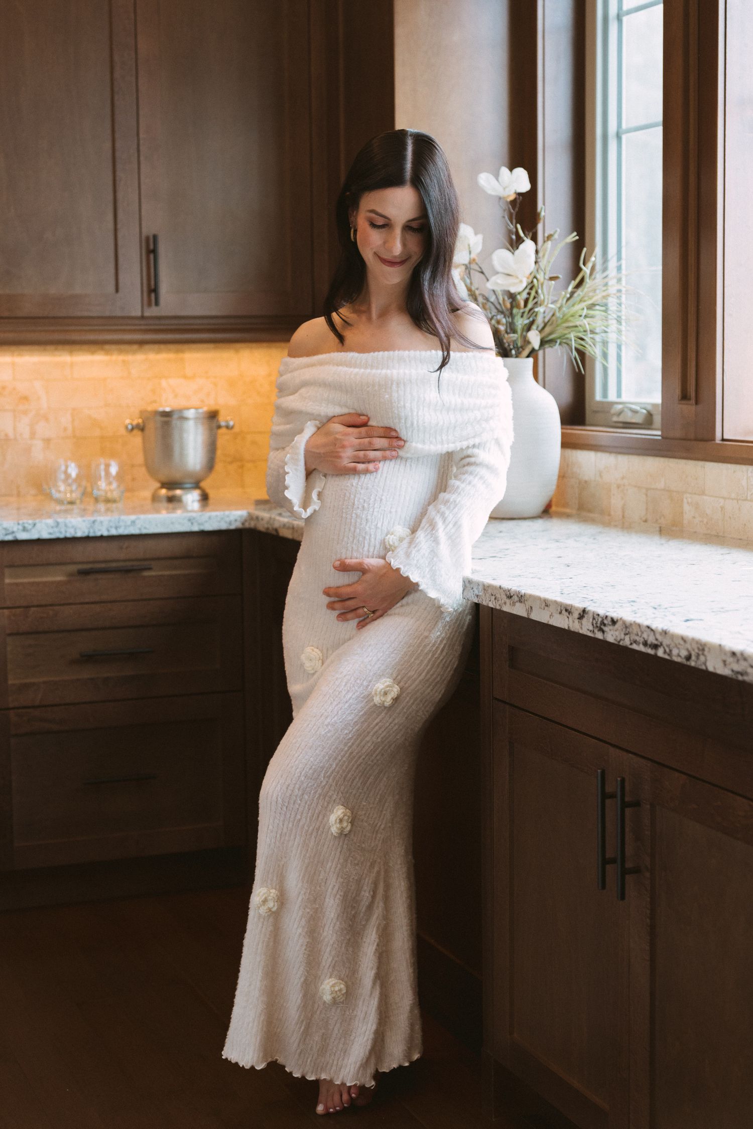 Pregnant woman in white floral knit off-shoulder dress cradling baby bump in warm wood-toned kitchen.