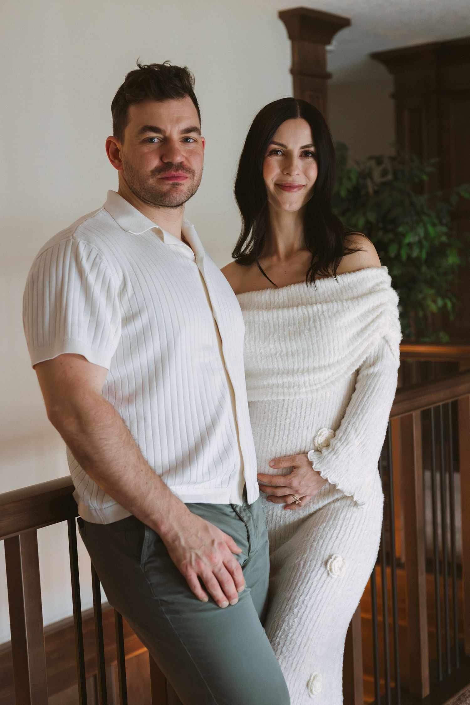 Stylish couple poses on indoor balcony, man in white ribbed shirt stands beside pregnant woman in cream knit dress.