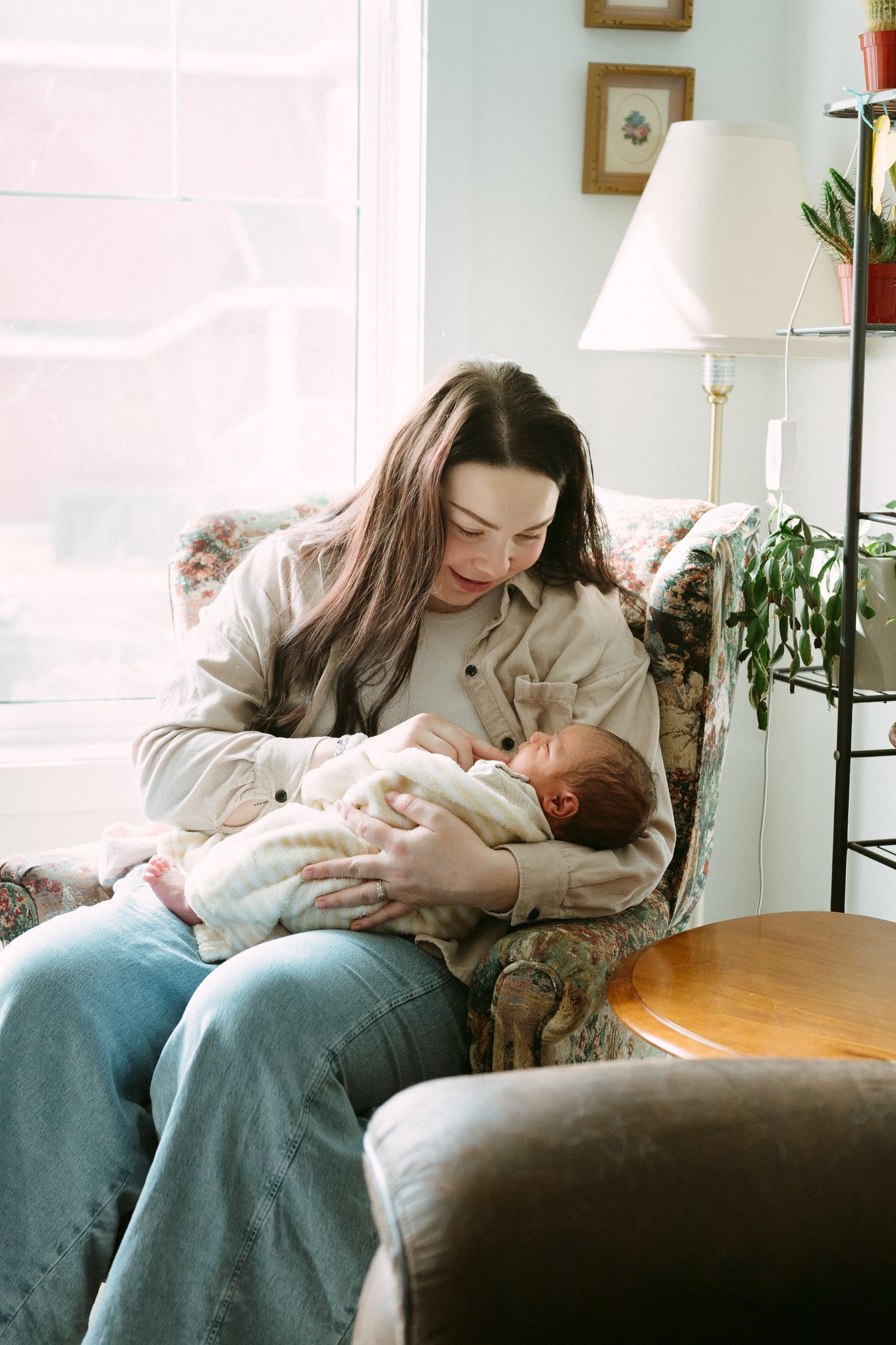 Young mother with long brown hair sits in floral armchair cradling newborn baby in cozy living room.