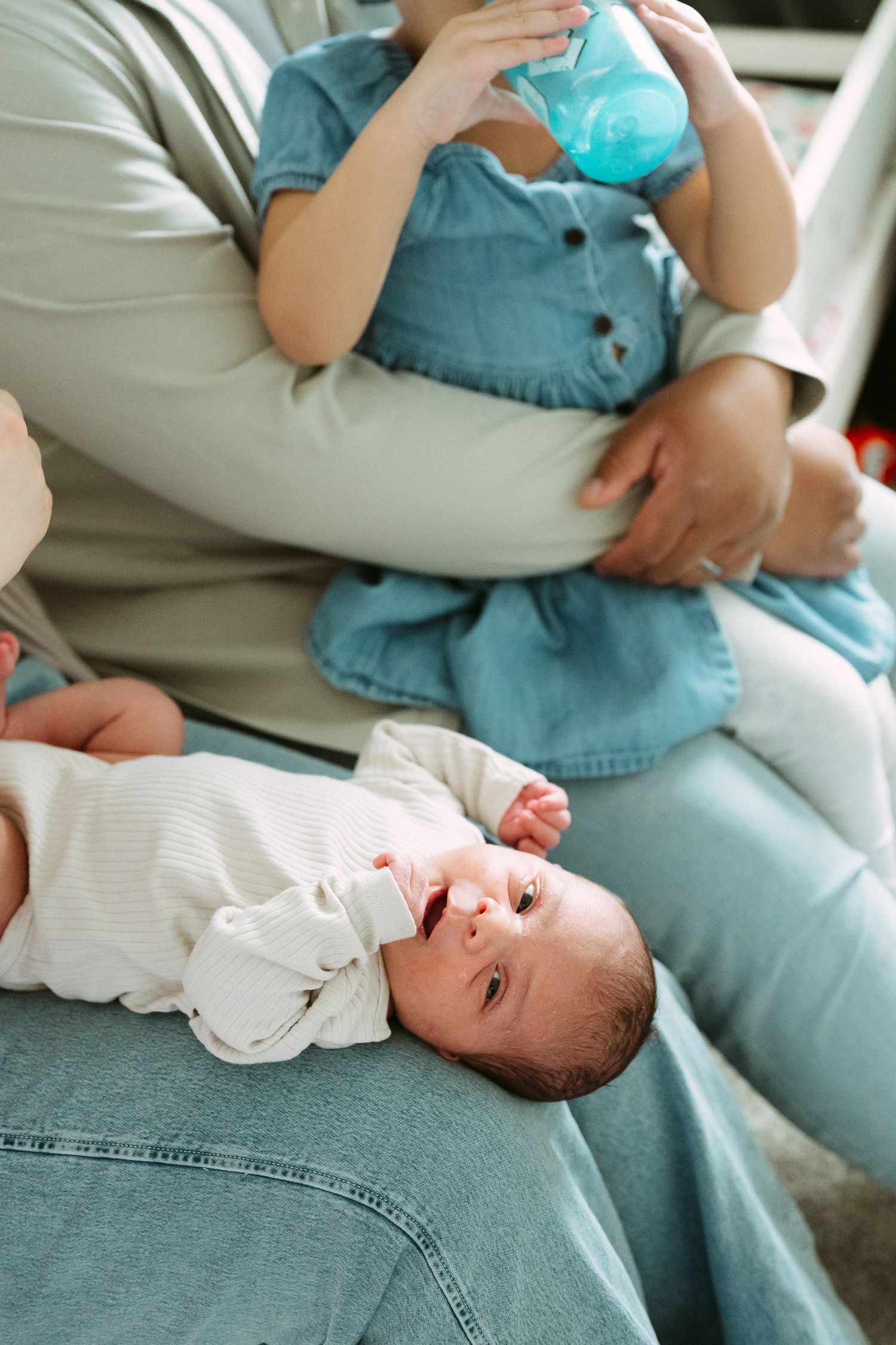 Mother sitting with toddler daughter drinking from sippy cup while newborn baby lies across her lap.