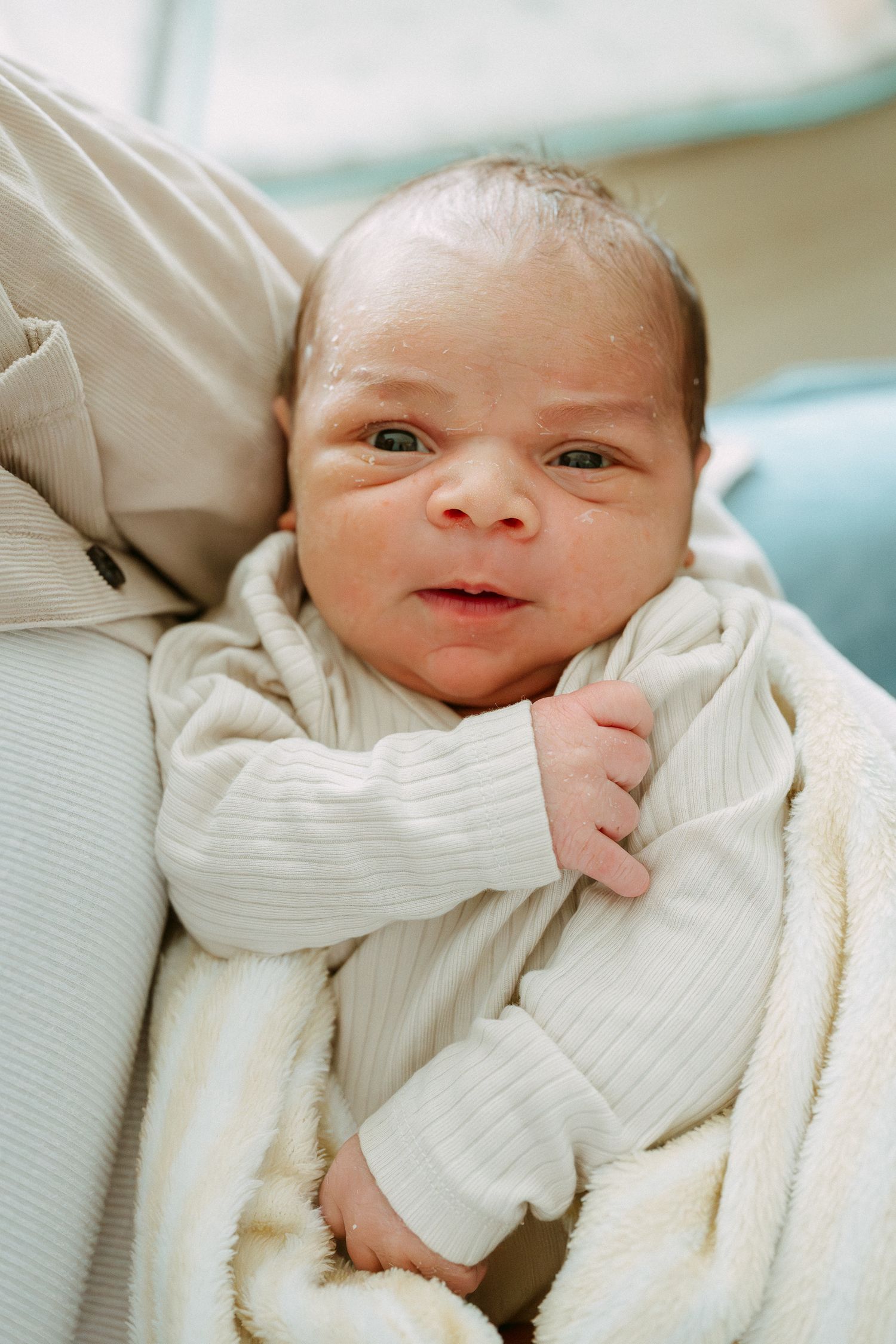 Newborn baby with dark hair wearing a white ribbed onesie, cradled in arms with a soft blanket.