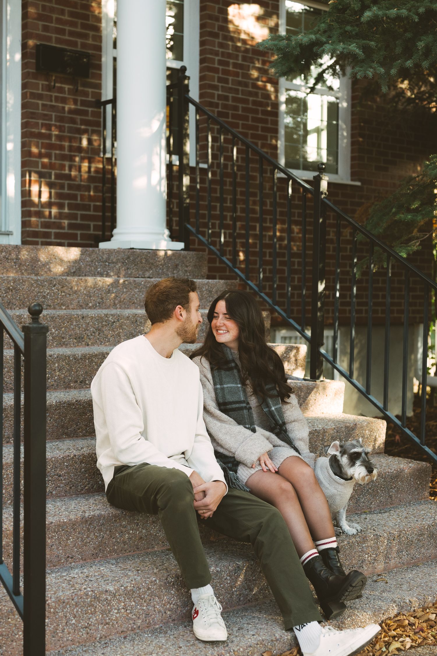 A couple sits together on outdoor stone steps with a small dog beside them on a sunny autumn day.