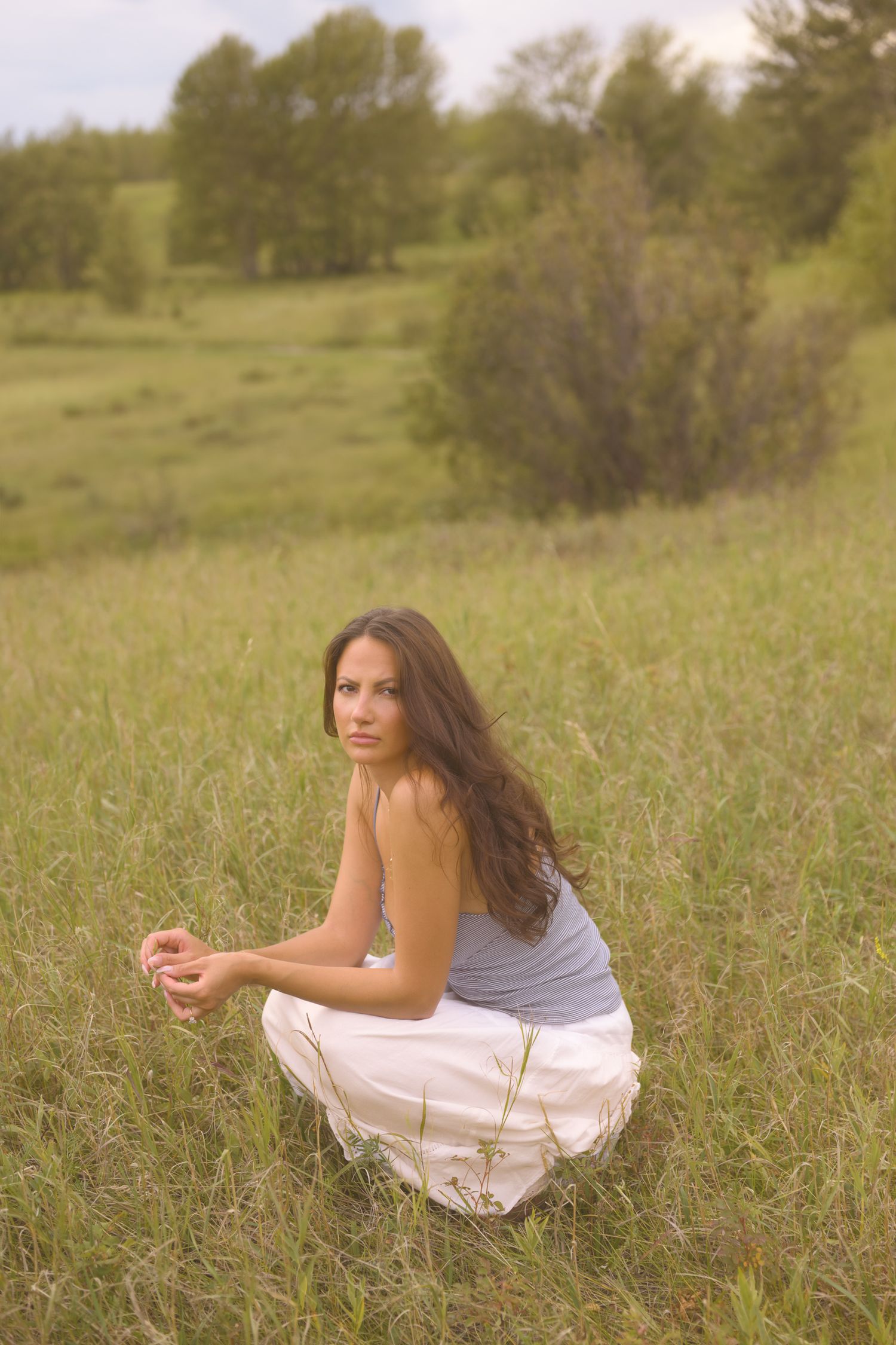 A figure in a white dress sits thoughtfully in a sunlit meadow with tall grass and trees in the background.