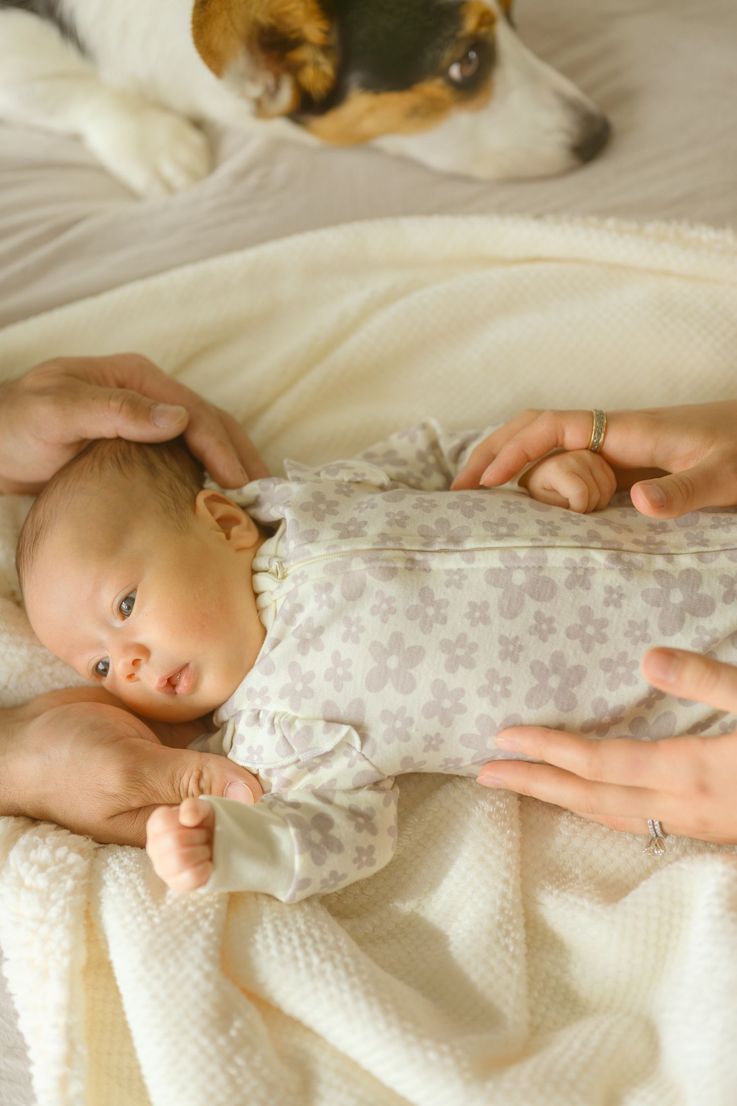 Infant wearing floral onesie lying on white bedding while hands gently hold them.