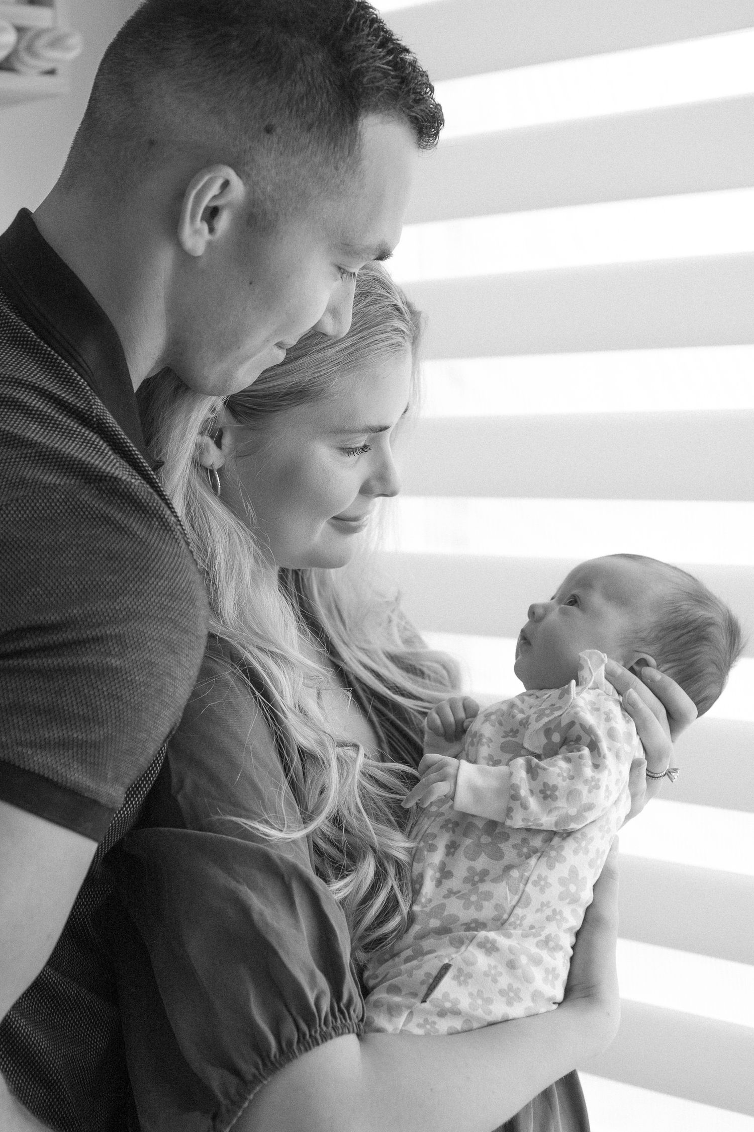 Tender family moment captured in soft window light showing parents bonding with their infant.