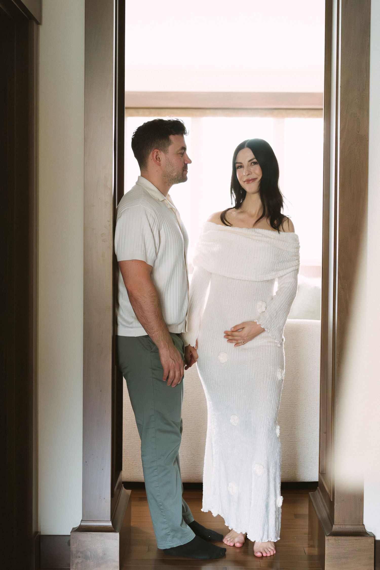 Expecting couple stands in a sunlit doorway, man in white shirt holds hands with pregnant woman in elegant white knit dress.
