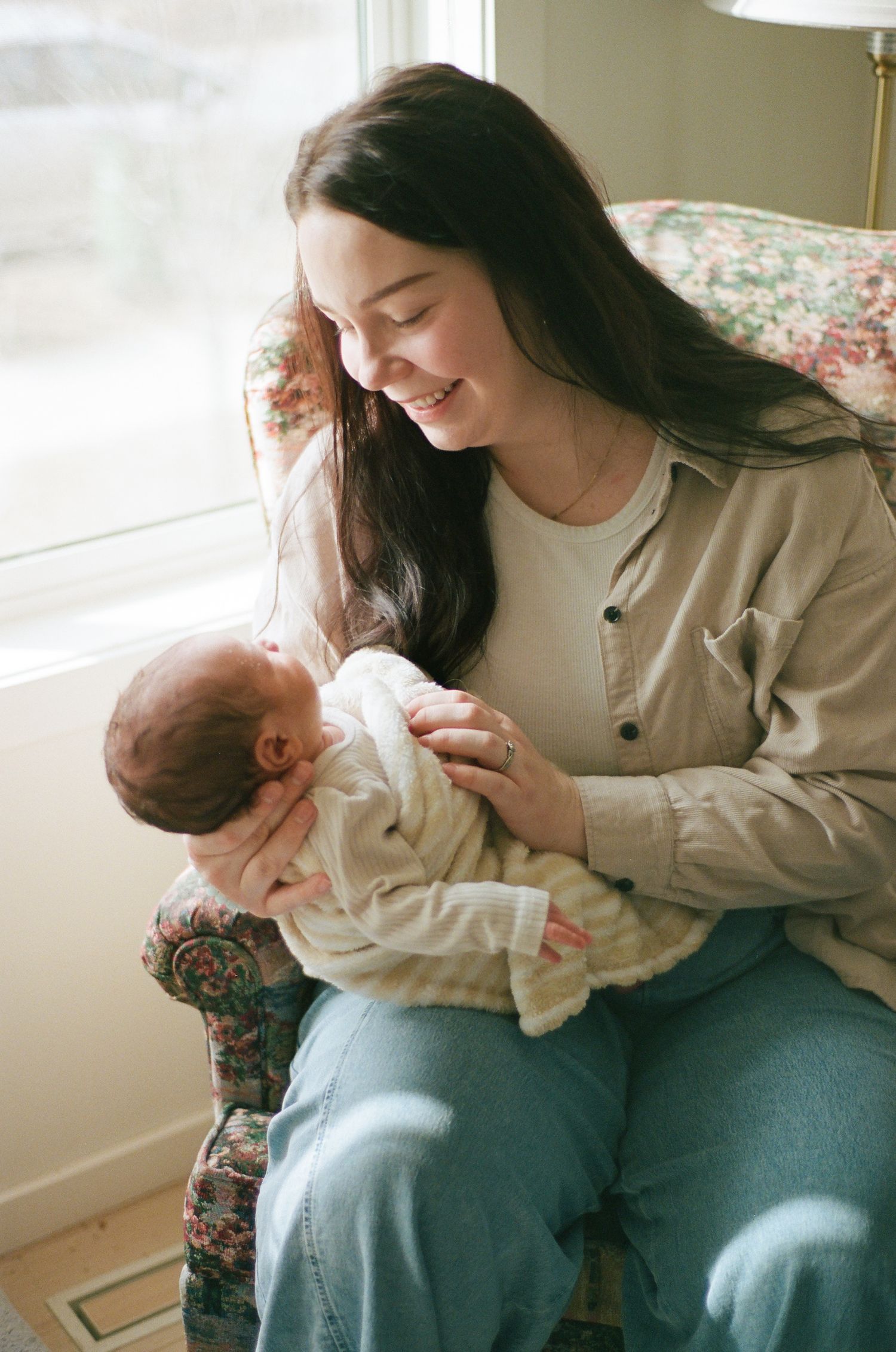 Young mother smiling warmly while cradling her newborn baby in a floral armchair by a bright window.