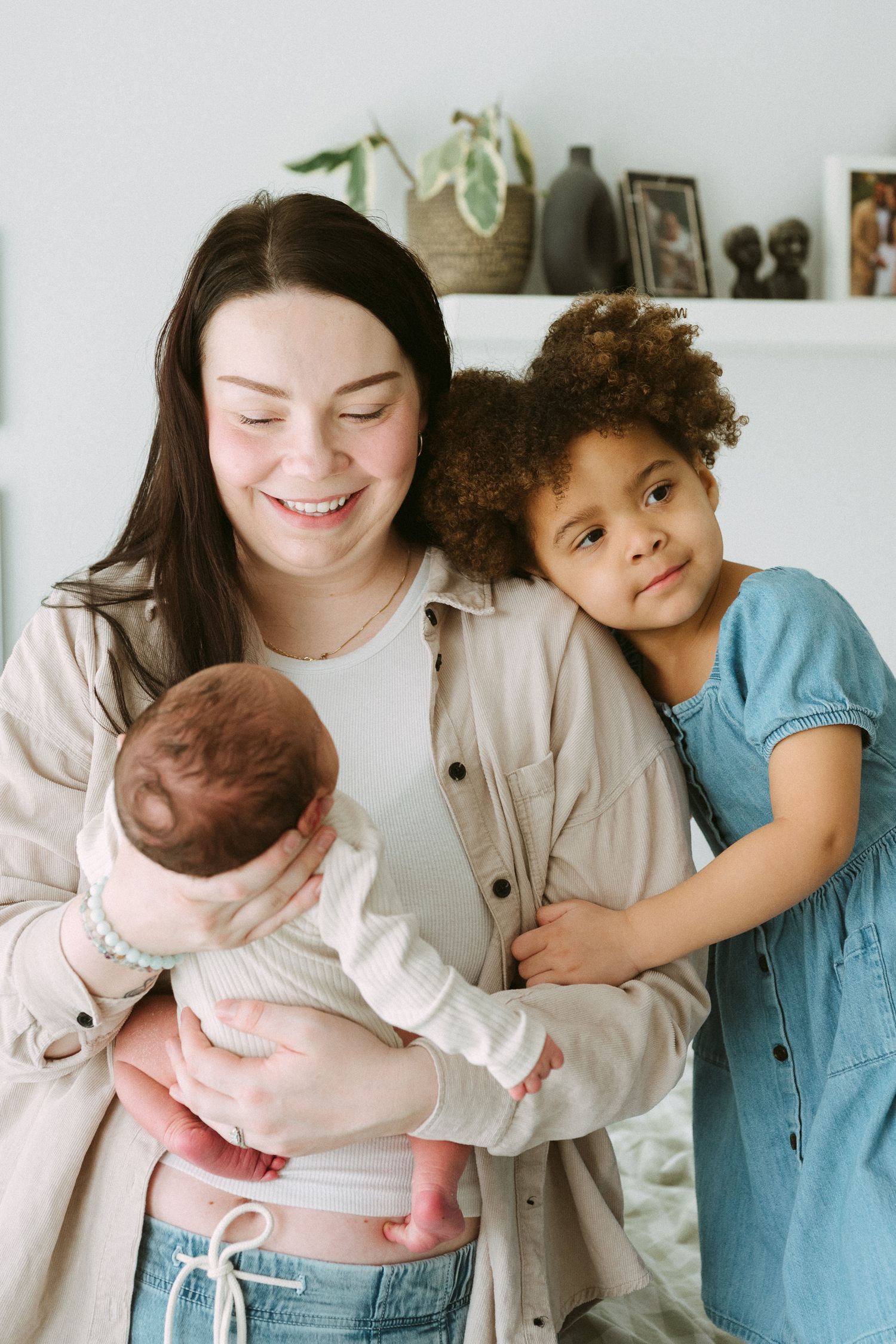 Smiling mother holding newborn baby while older curly-haired child hugs her in a bright home setting.