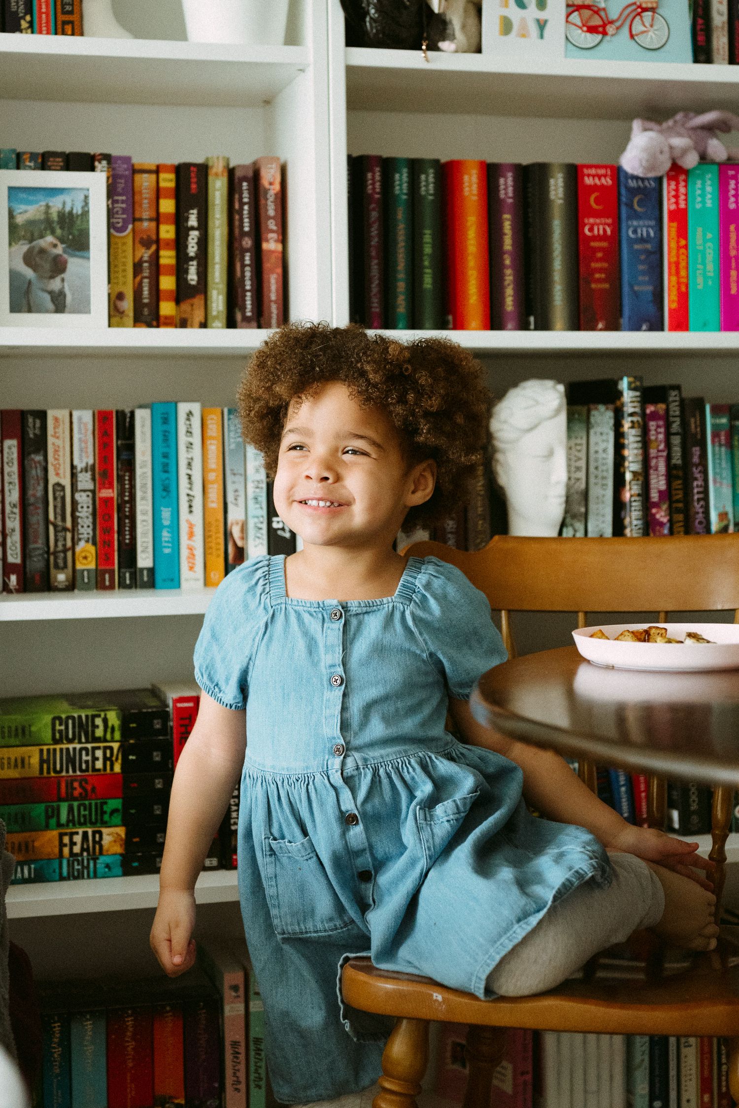 A young girl in a denim dress sits smiling in a wooden chair in front of a colorful bookshelf.