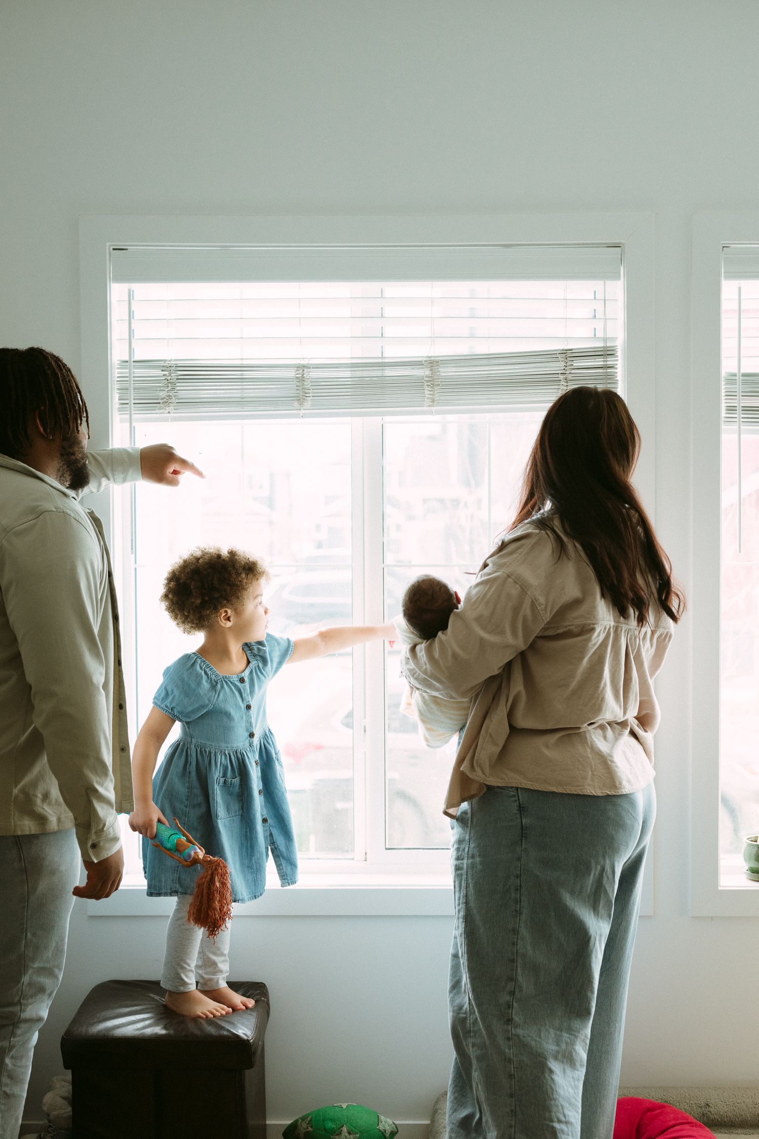 A multiracial family with two children points out the window together in a bright living room.