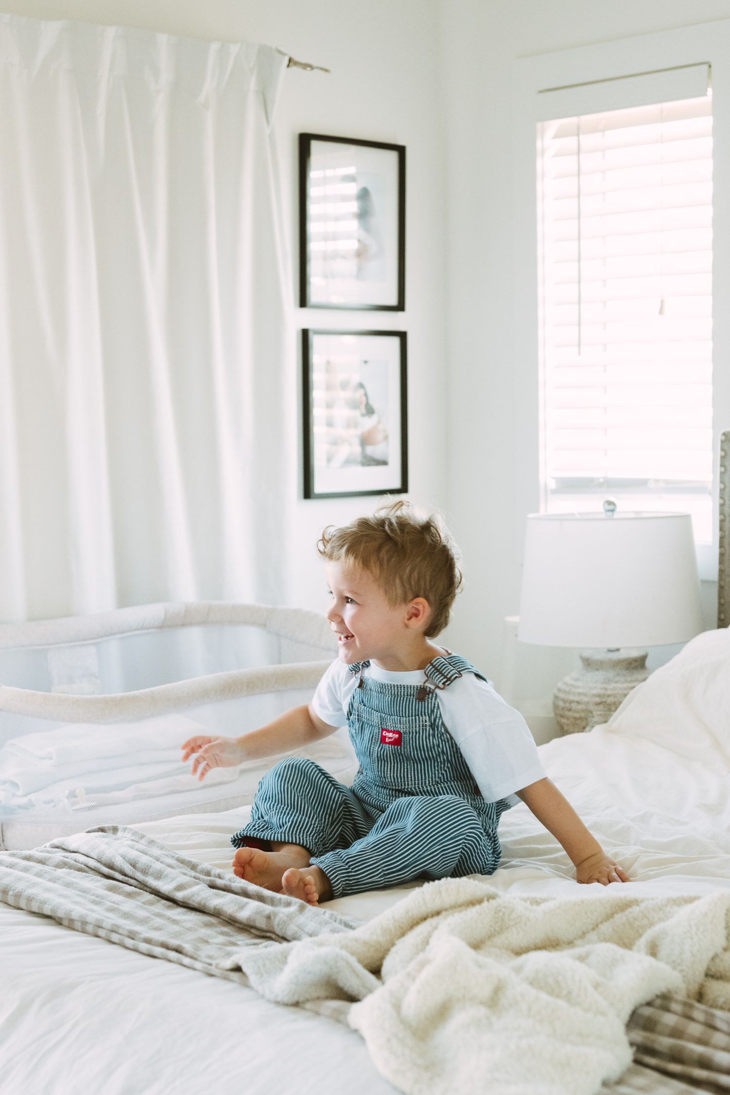 A small child in blue overalls sits on a white bed with framed artwork on the wall behind them.