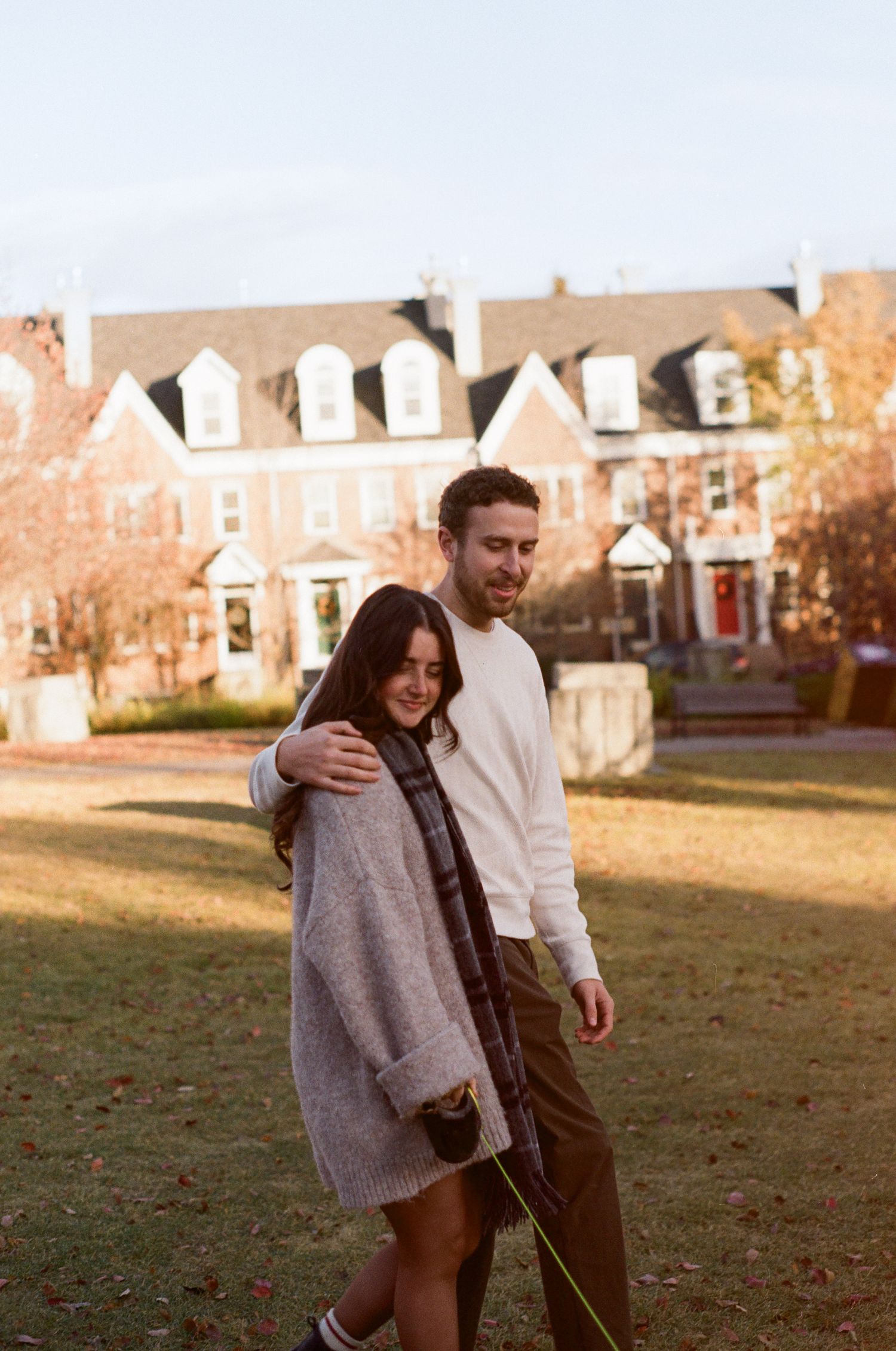Couple walking together on grass with brick townhouses in background during golden hour.