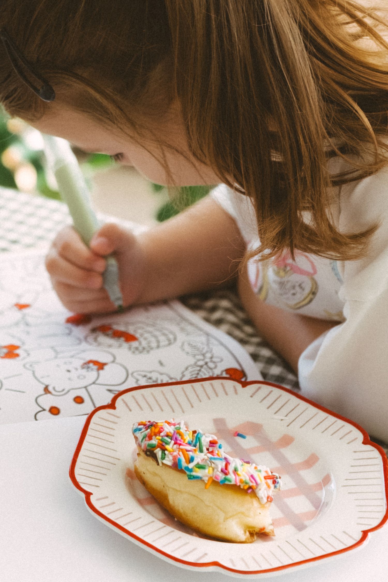 A colorful sprinkled donut on a red-trimmed plate sits beside someone drawing or coloring at a table.