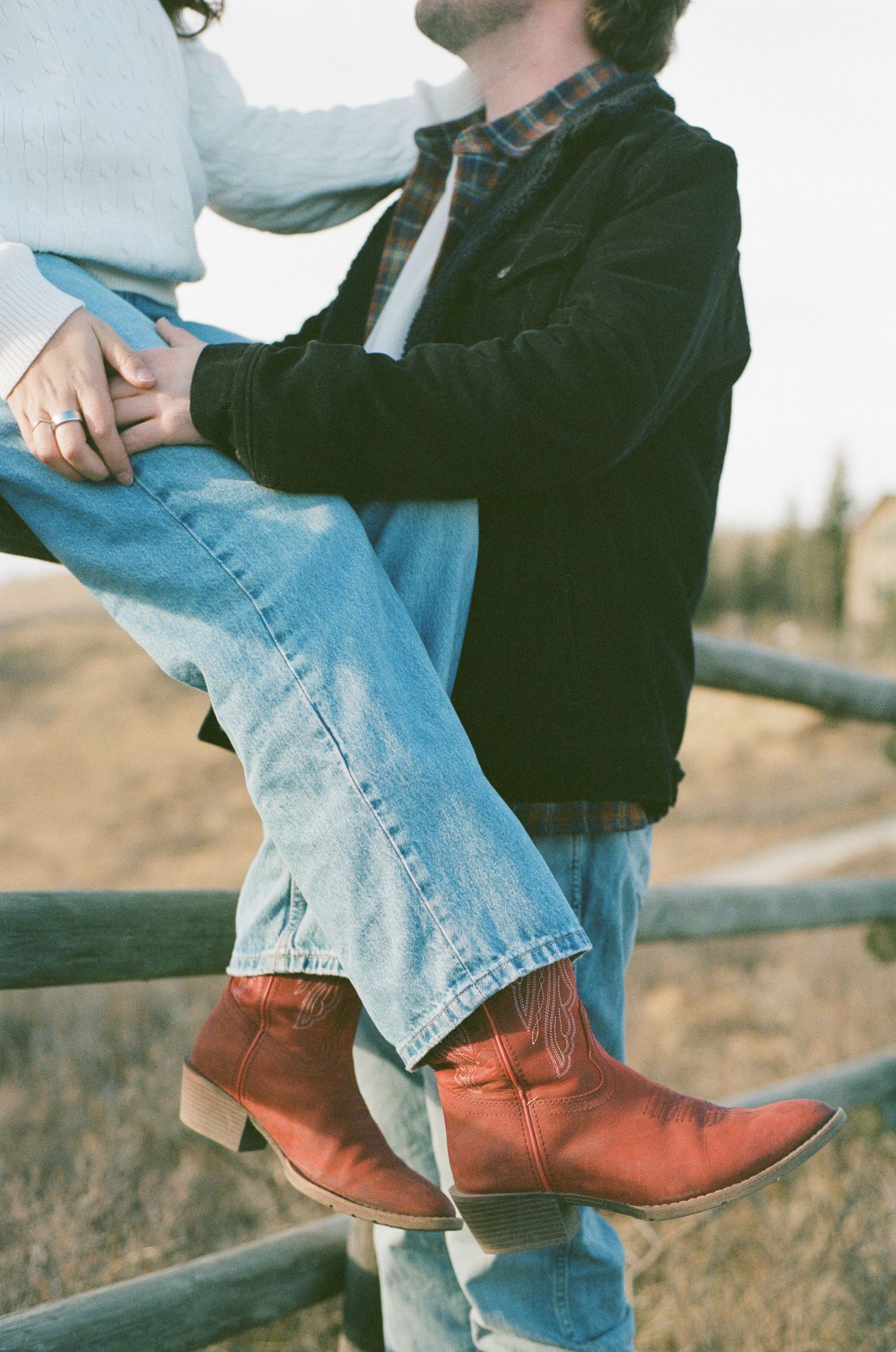 Person wearing blue jeans and brown leather boots sits casually on wooden fence in rural setting.