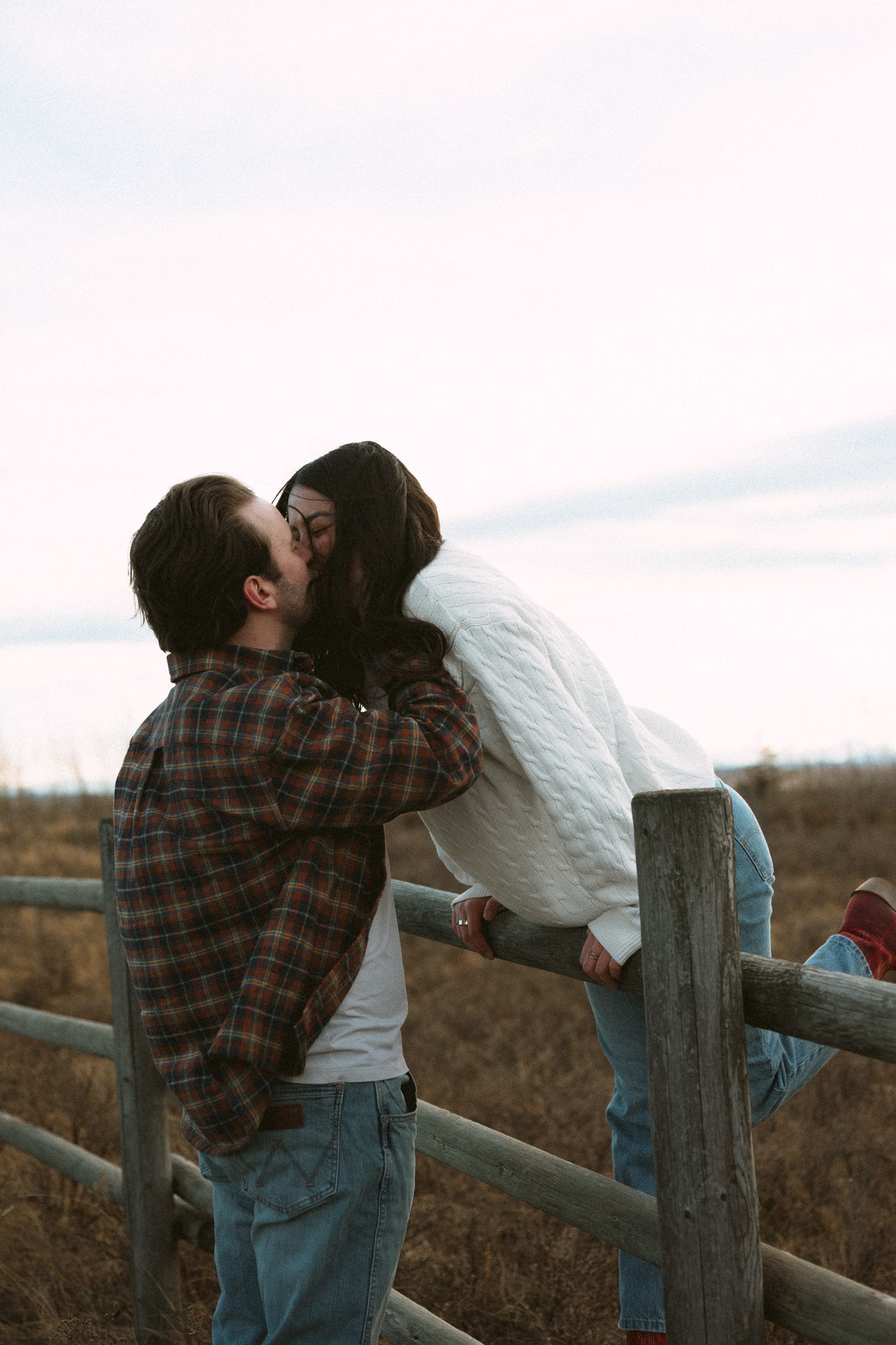 A couple in casual attire sharing a romantic moment while leaning against a wooden fence in a rural setting.