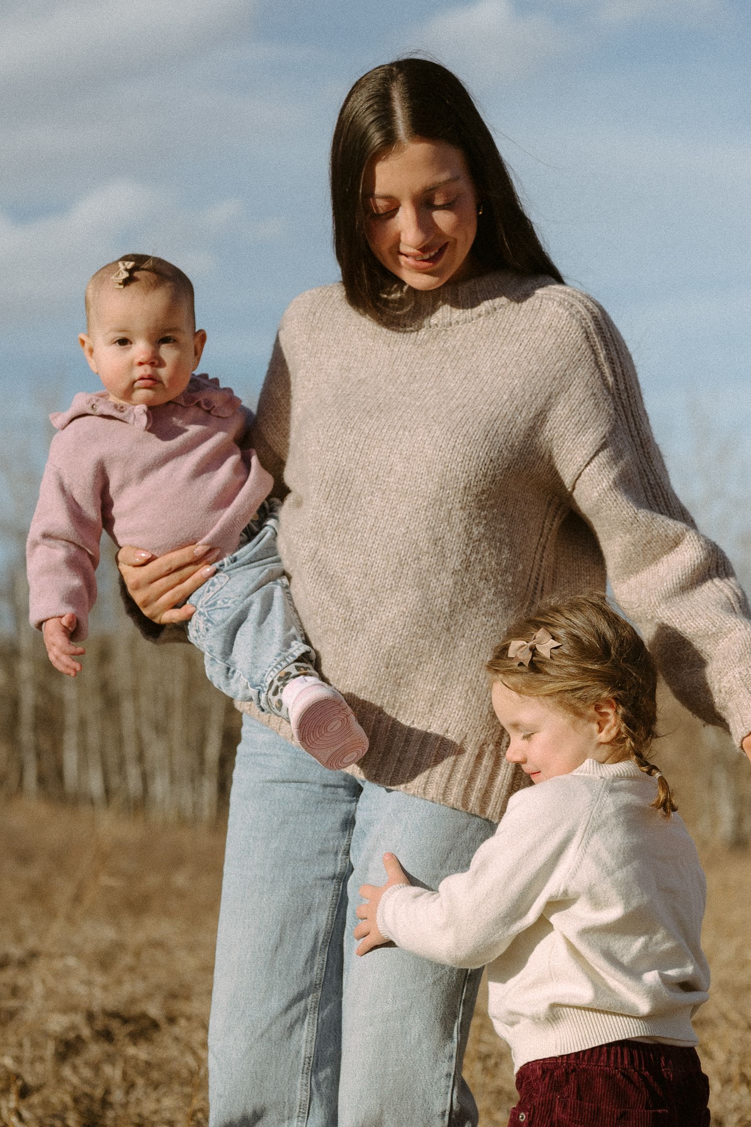 A tender family moment outdoors with a parent in beige sweater holding two young children in light colored clothing.
