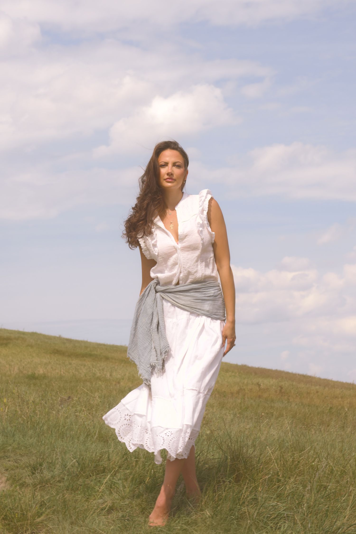 A figure in a white summer dress and gray sash stands barefoot in a meadow.