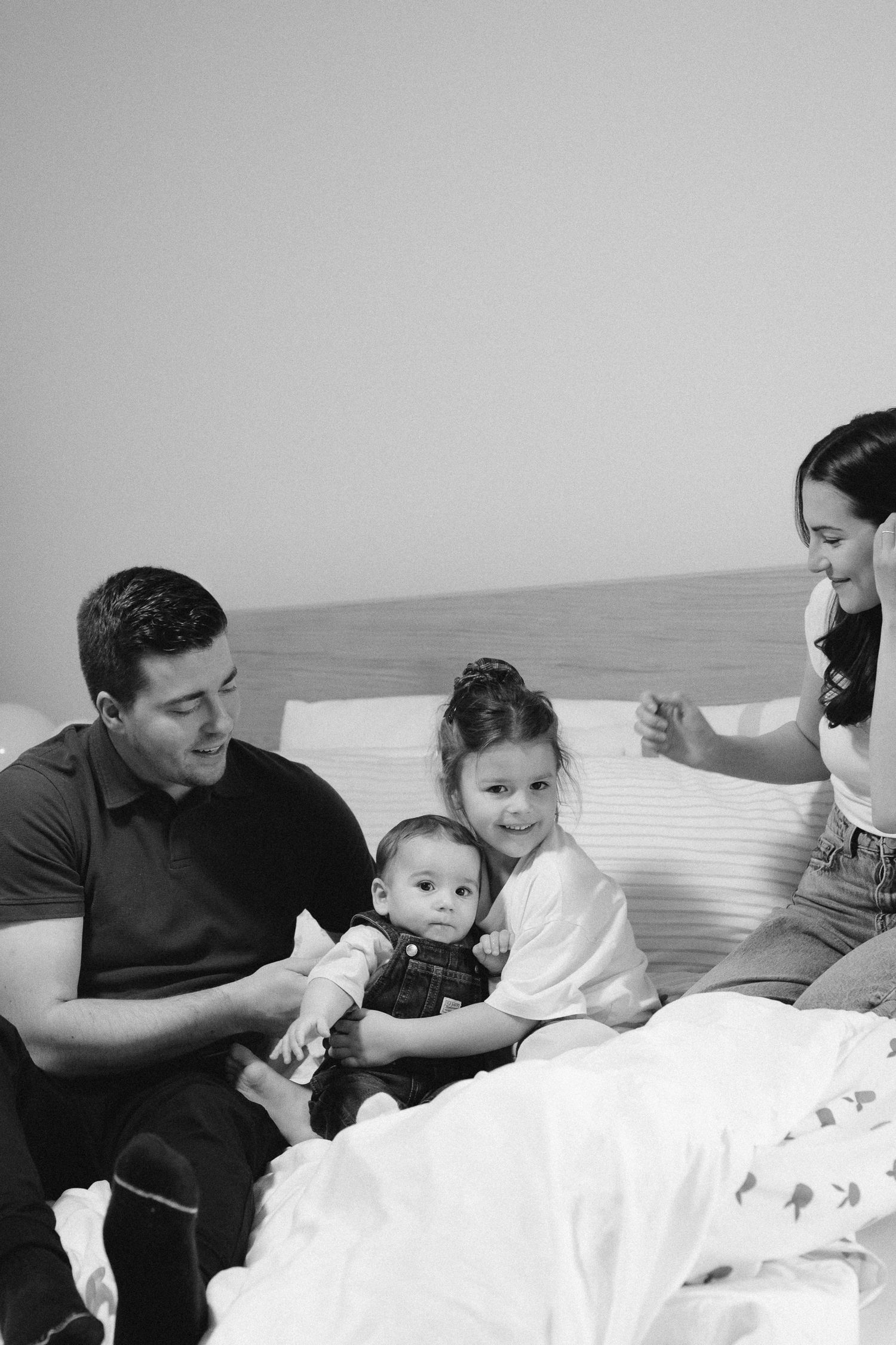 Family sits together on white bedding while sharing tender moments in black and white photo series.