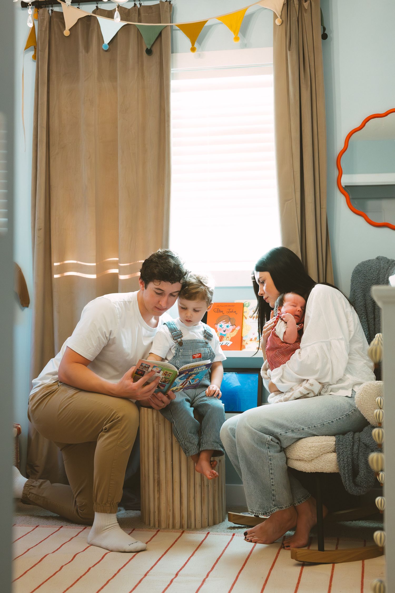 A family reads together in a cozy living room decorated with colorful bunting and soft curtains.