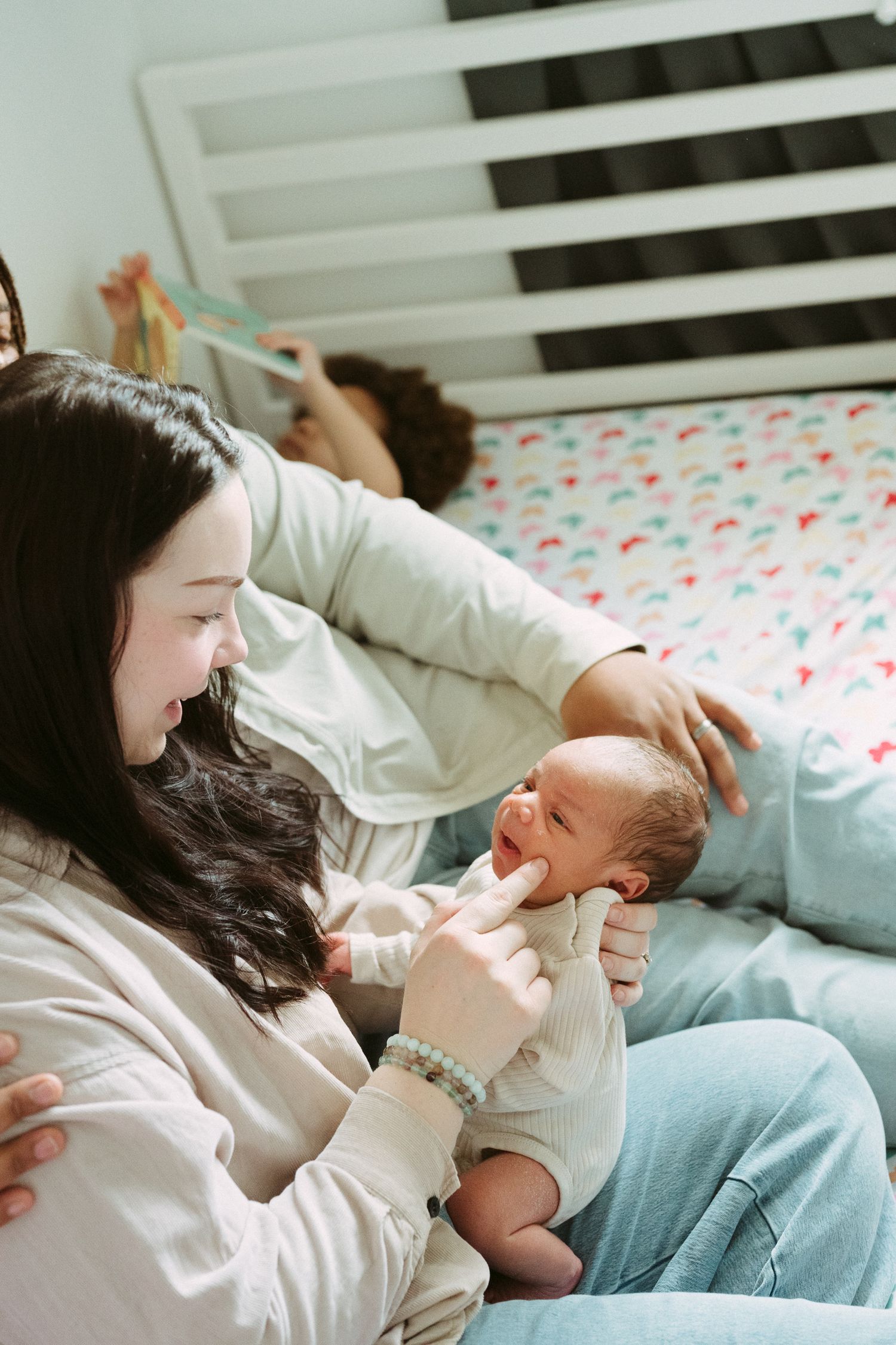 A mother smiles tenderly at her newborn baby while sitting on a bed, with another person resting in the background.