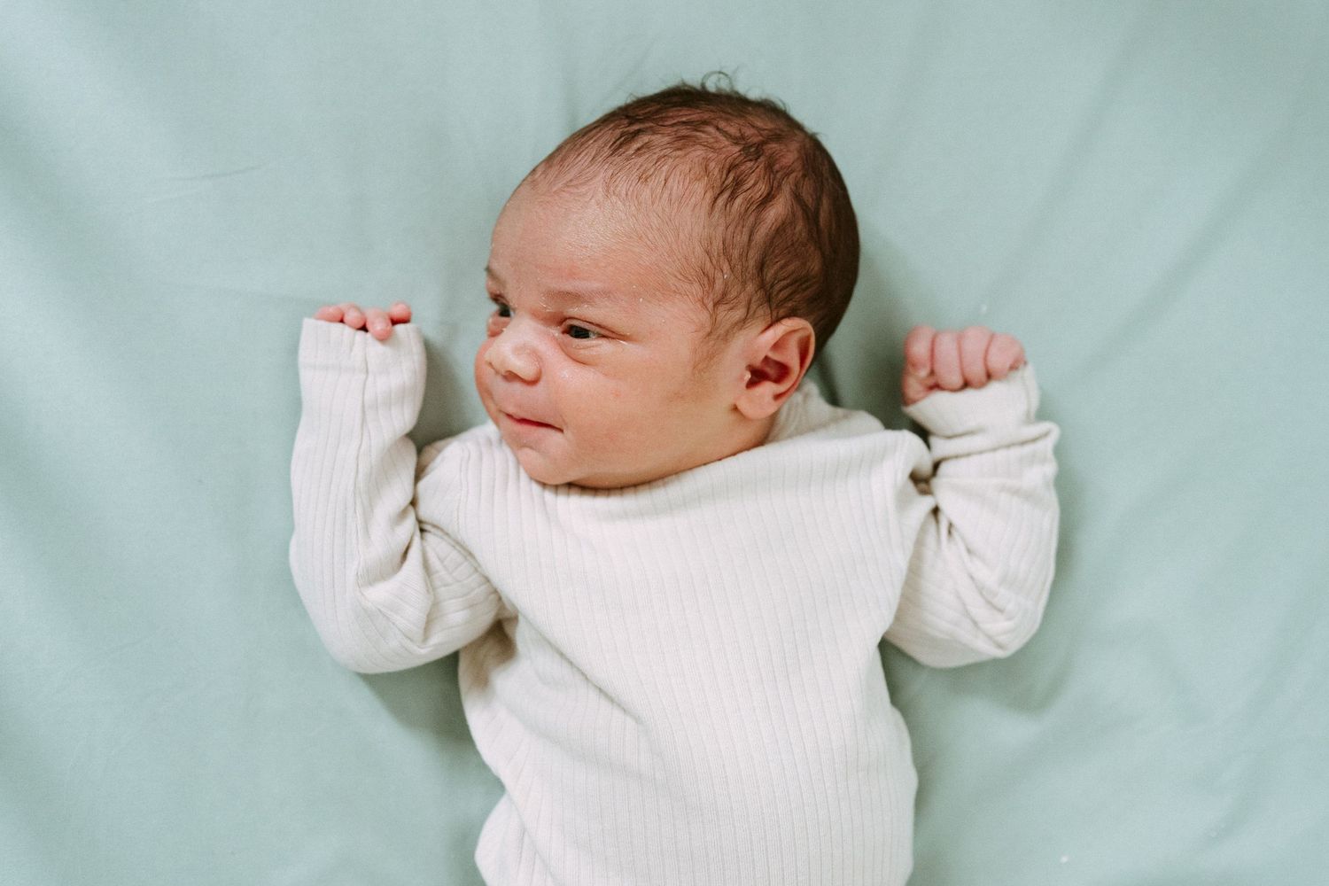 Newborn baby in white ribbed onesie lying on mint green blanket with arms raised, eyes slightly open.