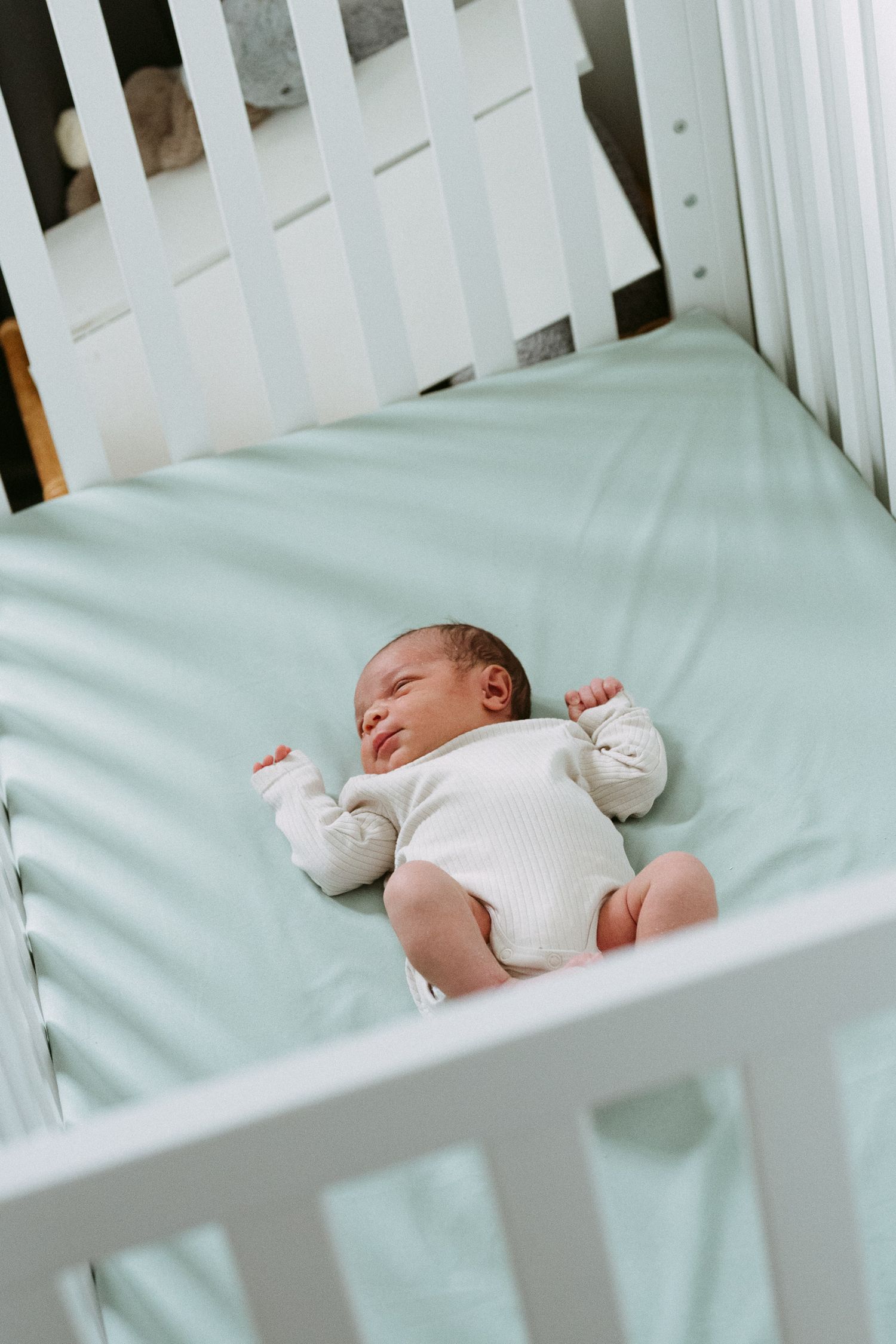 Newborn baby sleeping peacefully in a white crib on a mint green sheet wearing a white onesie.