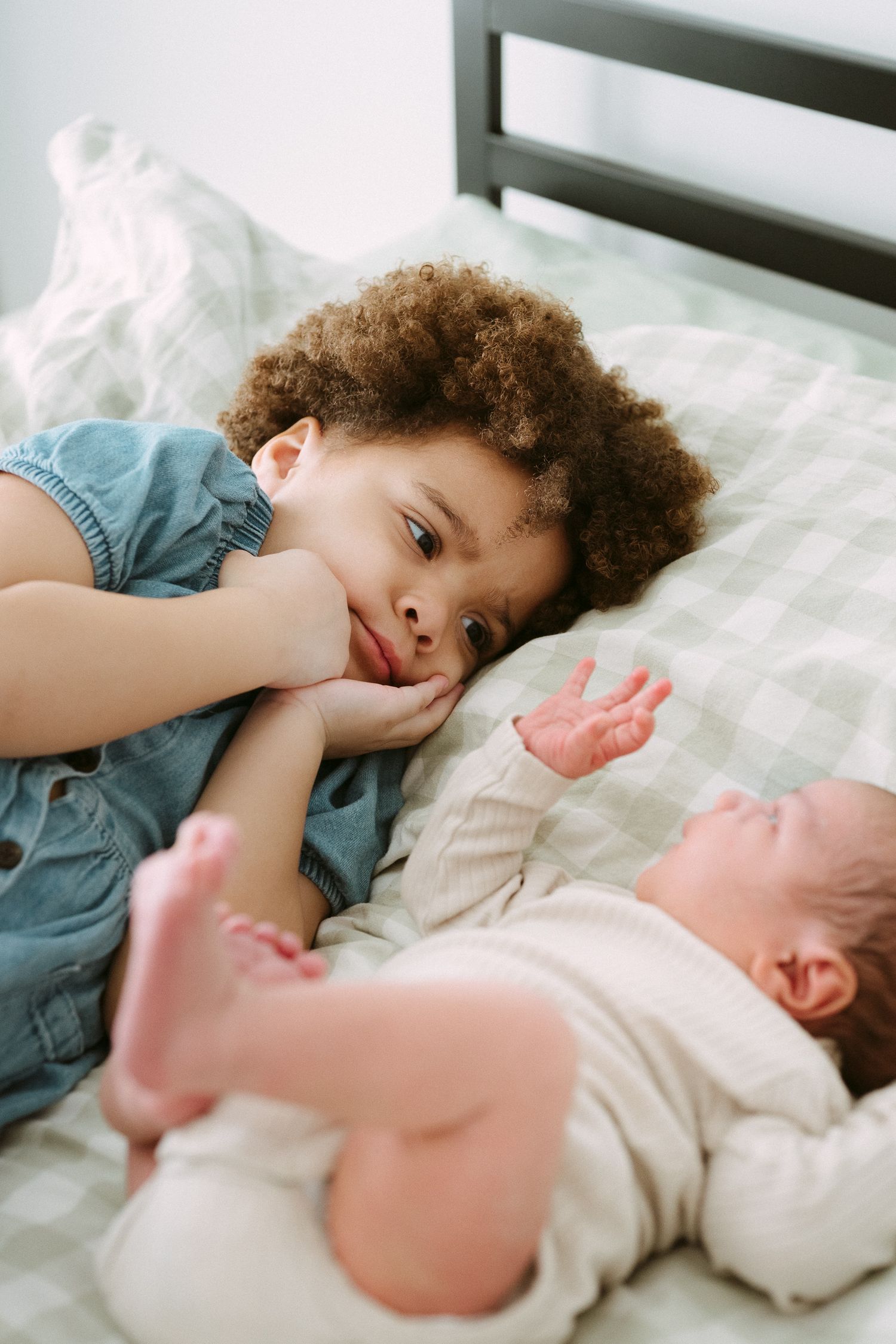 A curly-haired child in a denim dress lies on a bed gazing lovingly at a sleeping newborn baby sibling.