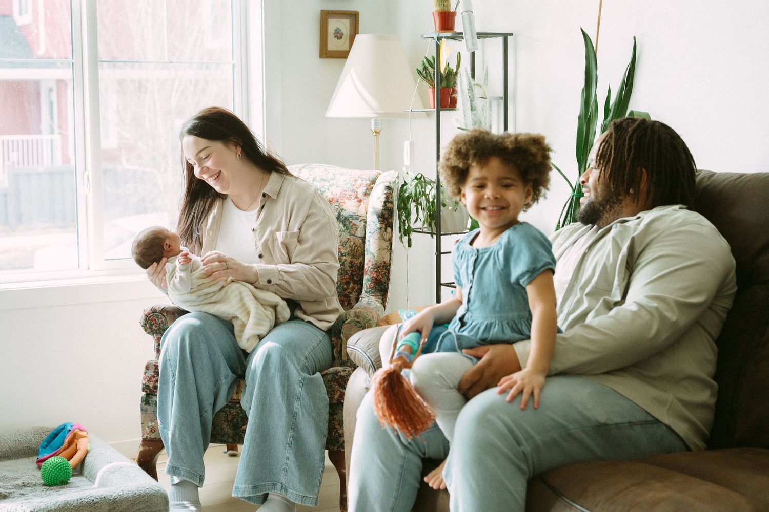 A young family relaxes on a couch, with a mother nursing a newborn and a father sitting with their curly-haired toddler.