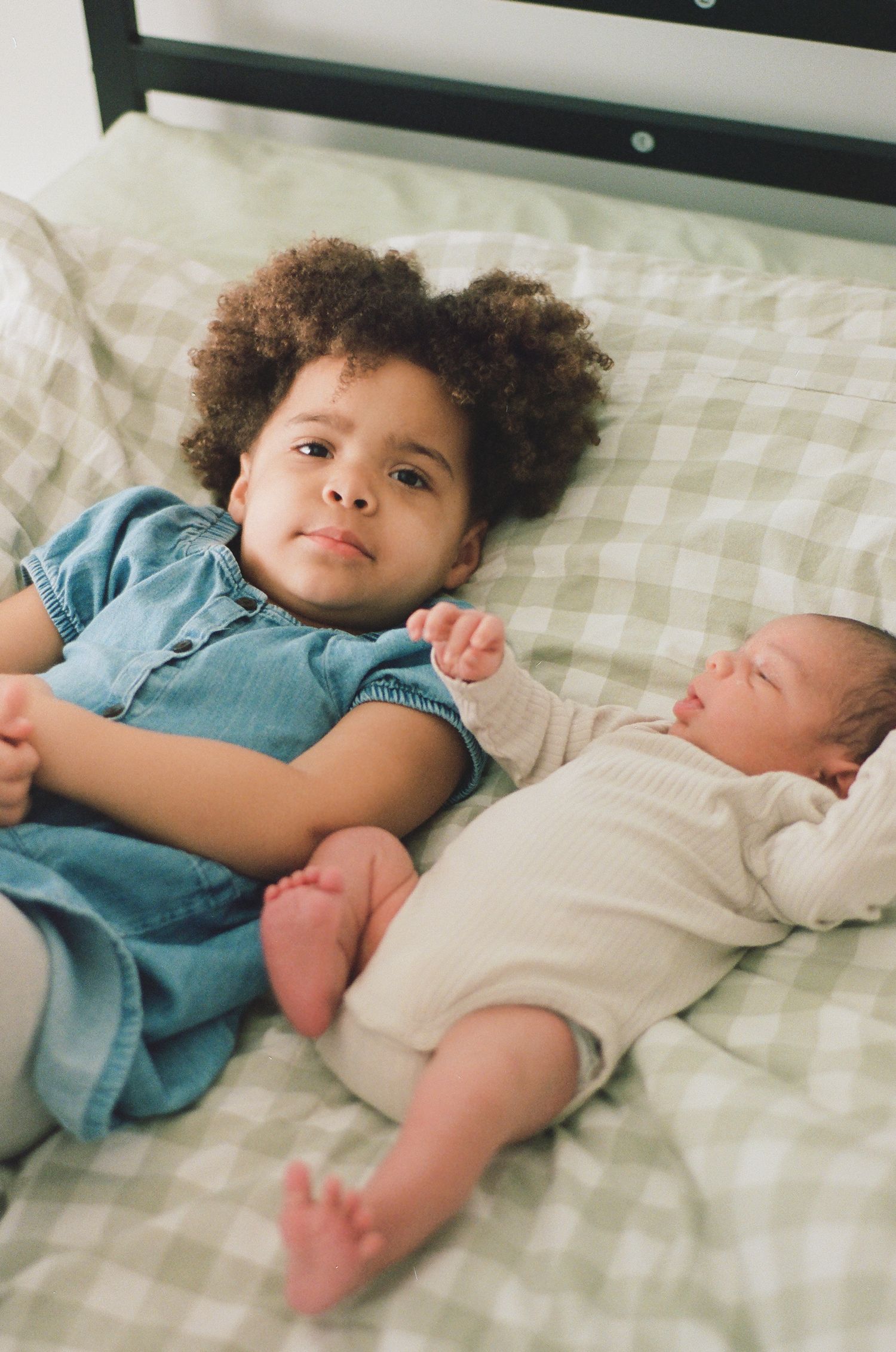 A toddler with curly hair in a blue dress lies beside a sleeping newborn baby on a green gingham bed.