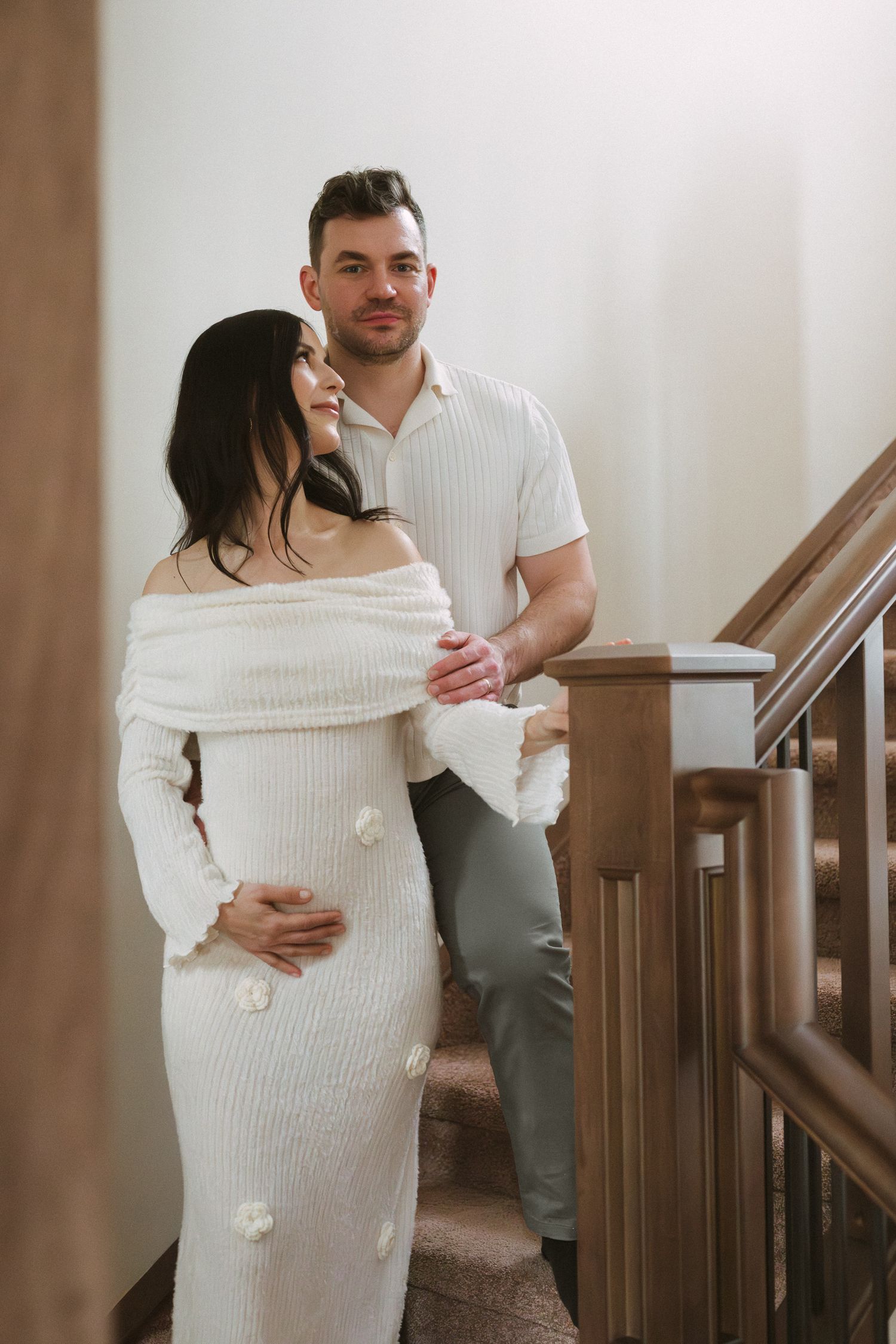 Couple on wooden staircase, man in white ribbed shirt embraces pregnant woman in cream off-shoulder knit maternity dress.