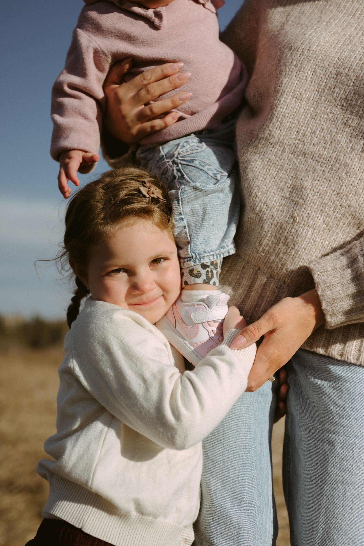 A young child in a white sweater hugs a parent's leg outdoors during a sunny afternoon.