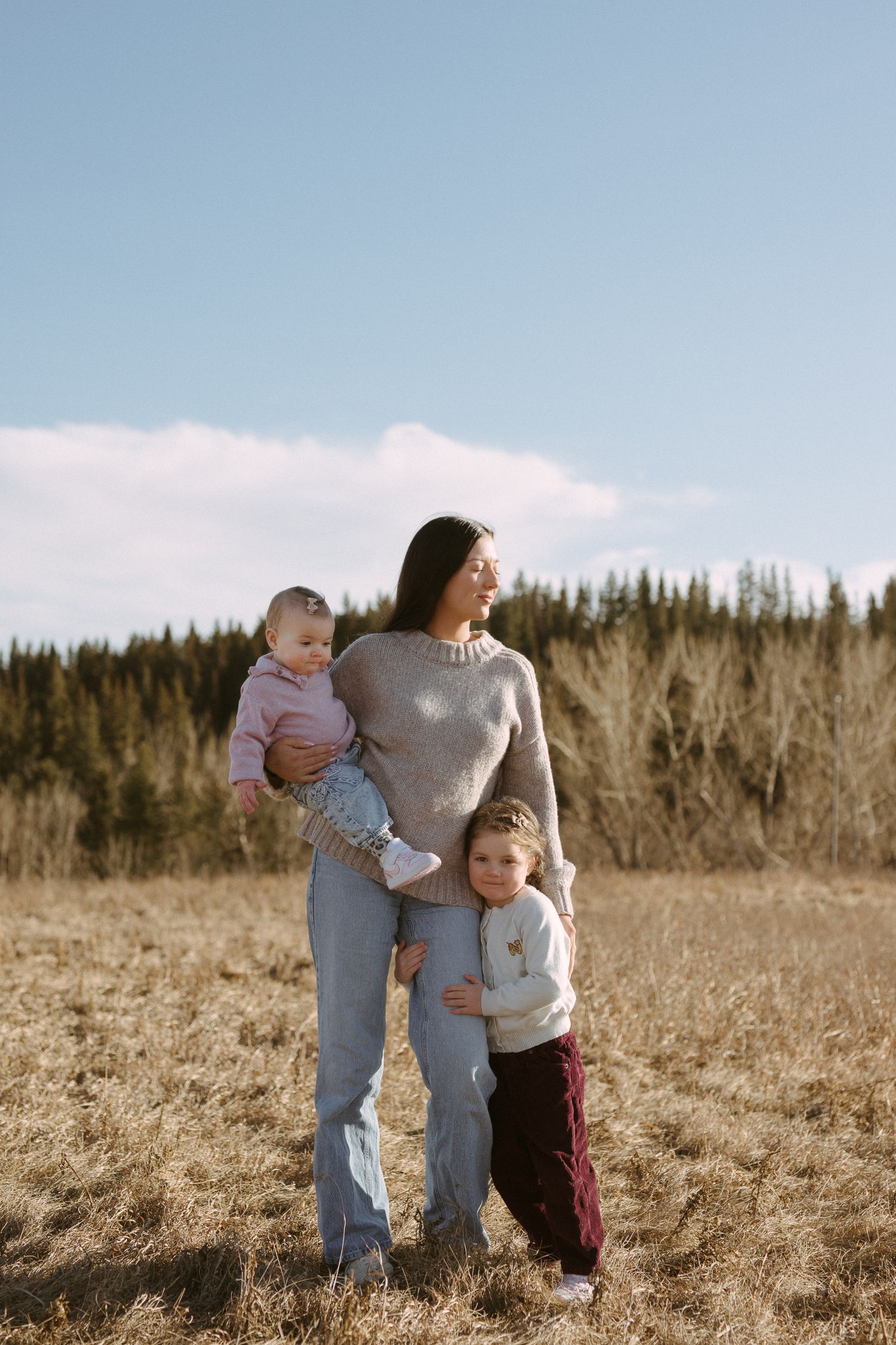 A parent and two children stand together in a sunlit field with evergreen trees in the background.