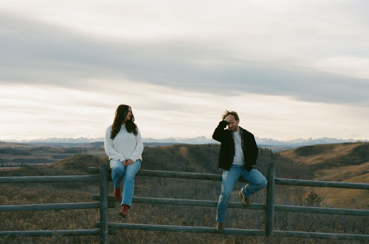 Two people sit on a wooden fence overlooking a scenic autumn landscape at sunset.