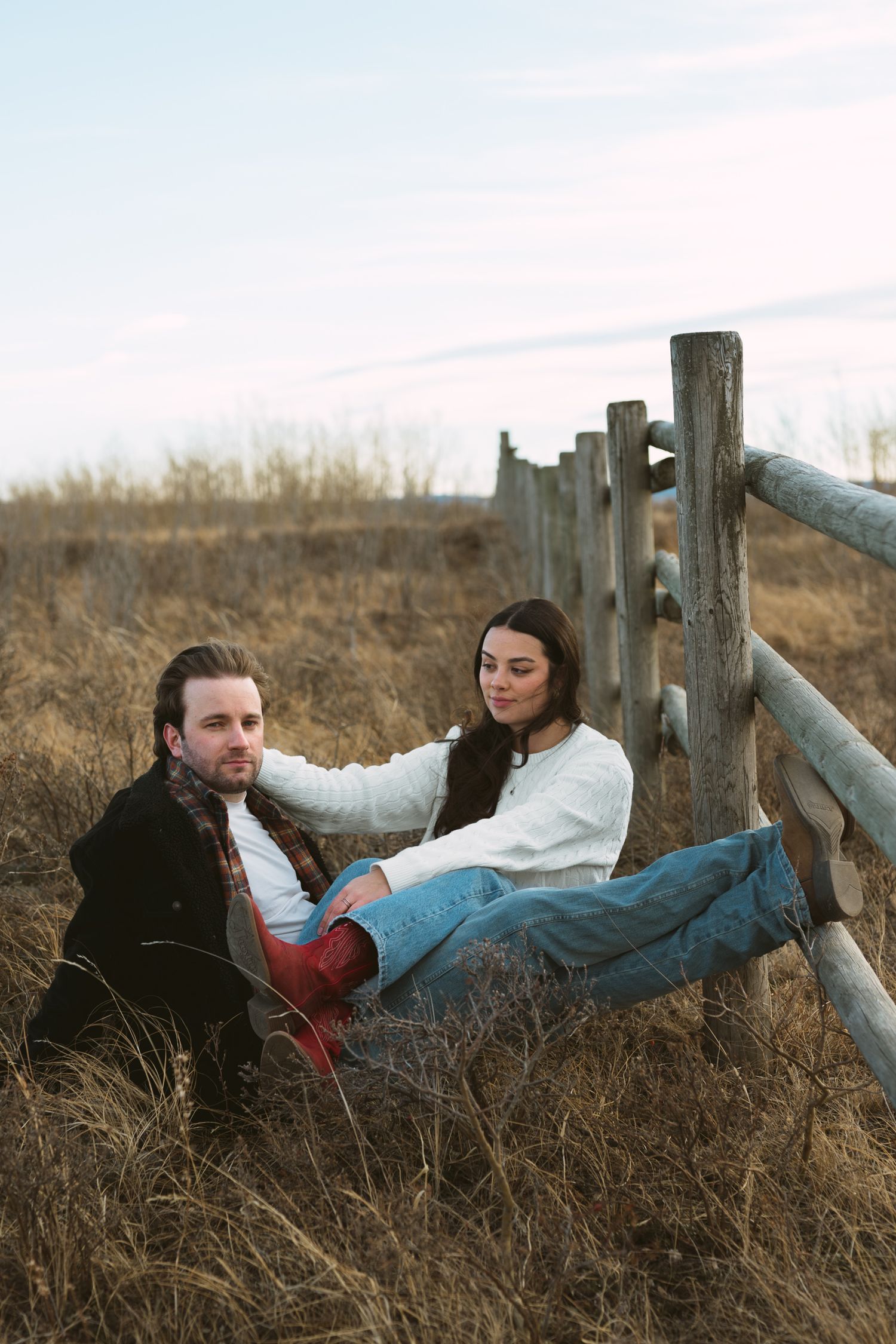 Couple relaxing by rustic wooden fence in autumn field during intimate photoshoot.