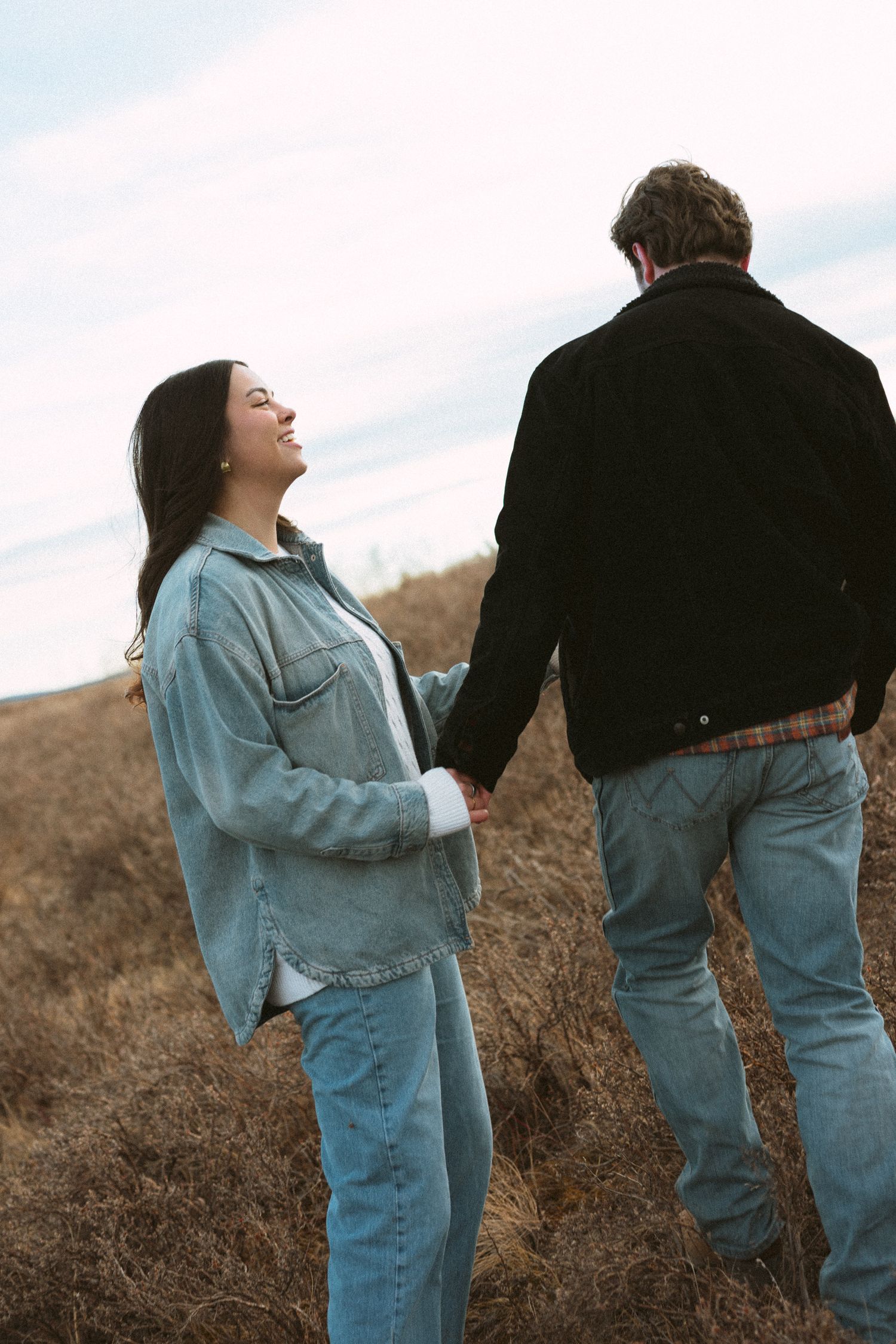 Two figures in casual denim attire climb up a brown grassy slope together.