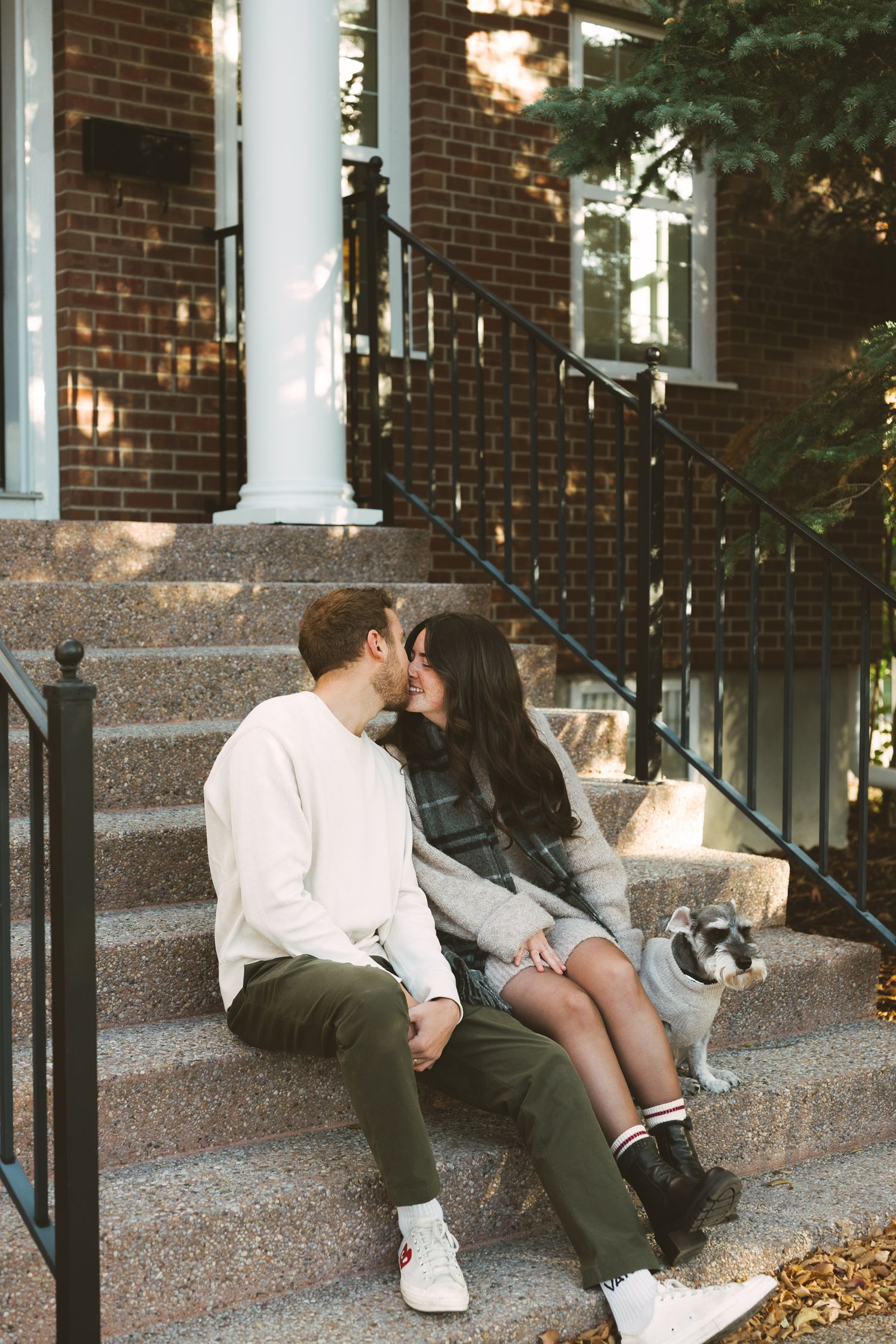 A couple sits intimately on concrete steps outside a brick building on an autumn day.
