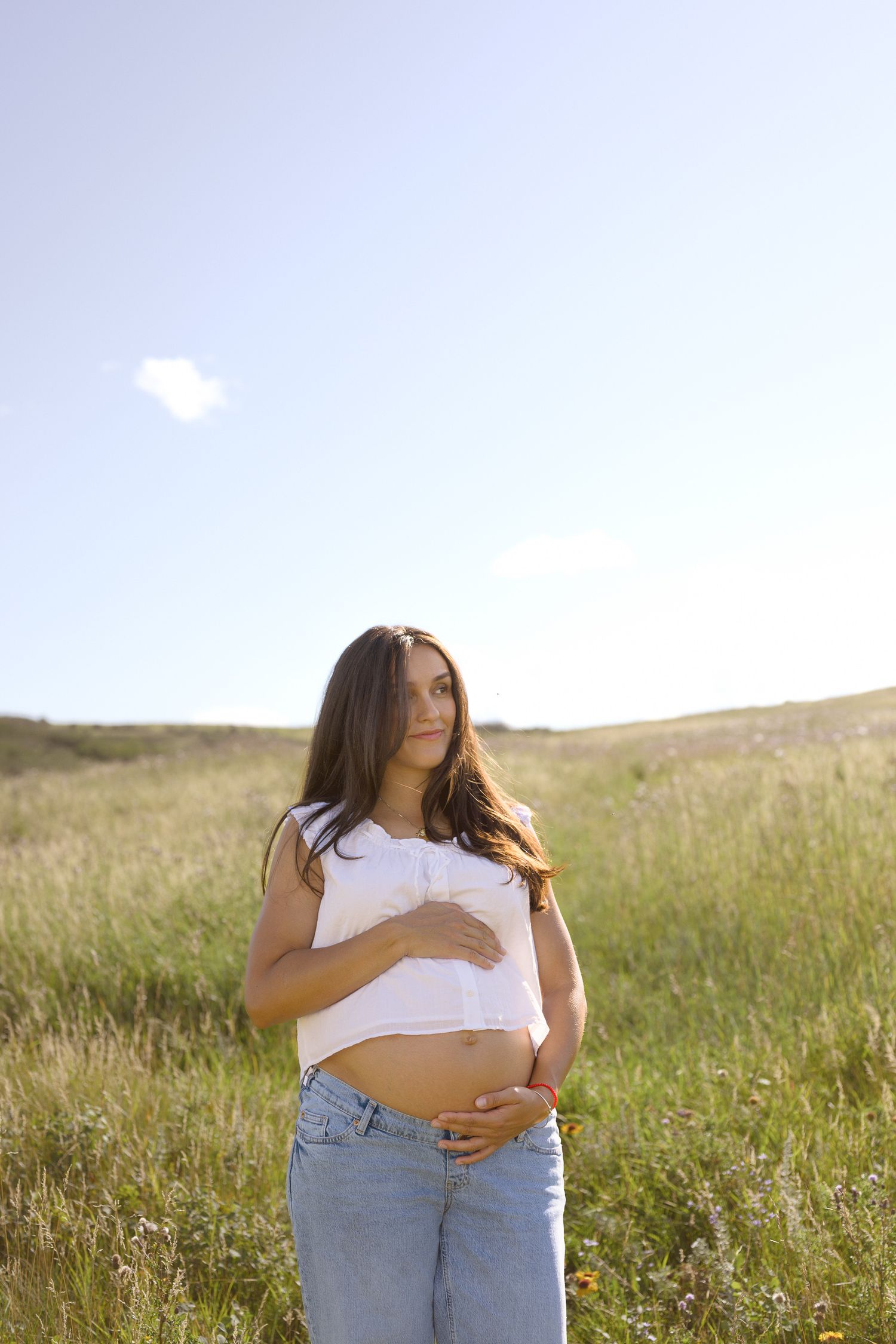 Woman in denim and white top embraces herself while standing in a sunlit meadow.