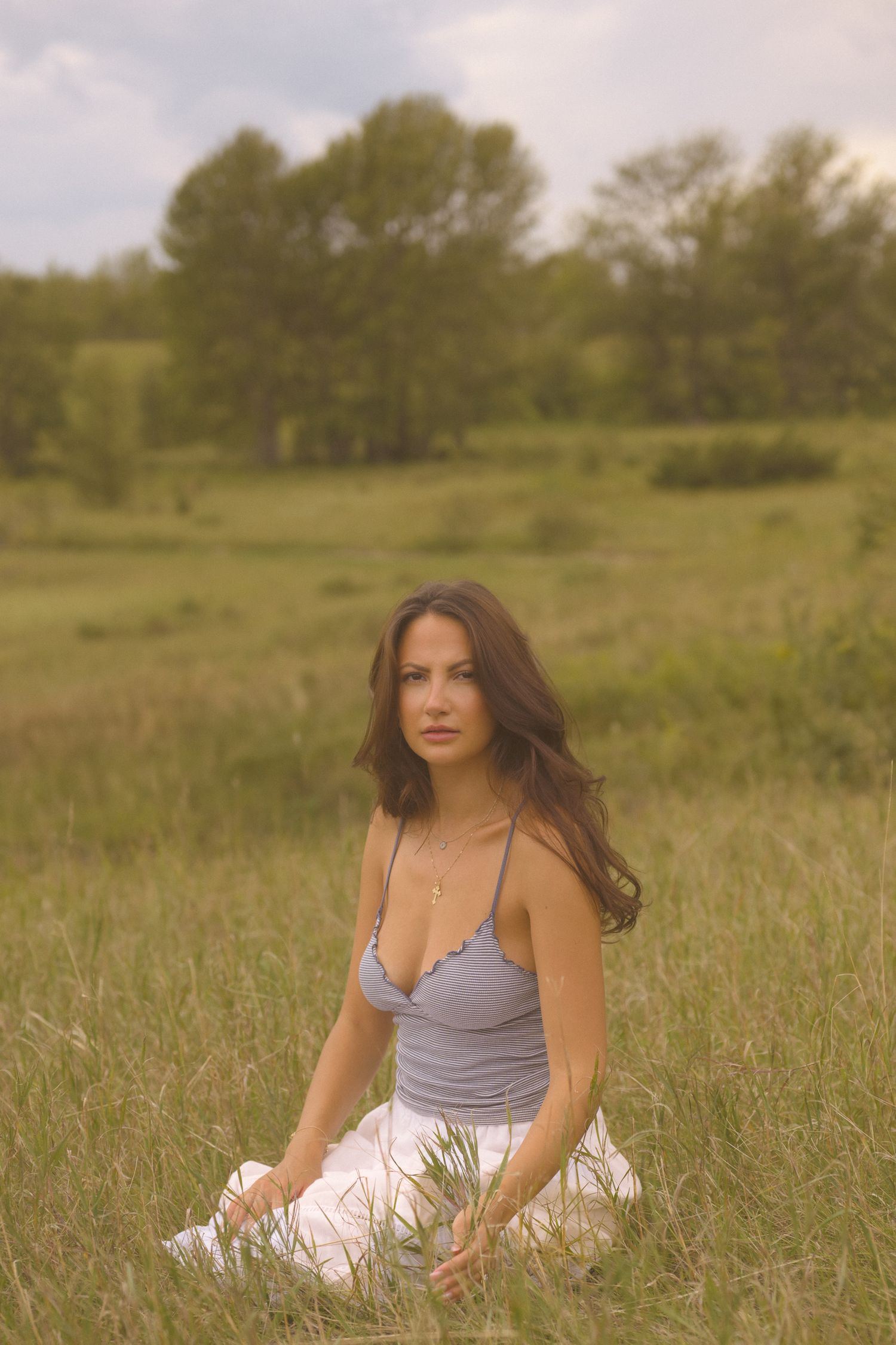 Person in striped top relaxes in a sunny meadow with tall grass and trees in the background.