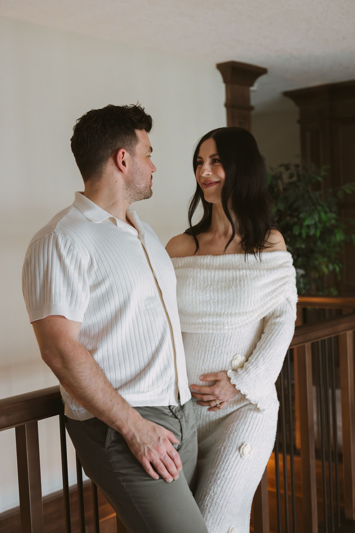 Couple shares tender moment on indoor balcony, woman in white off-shoulder maternity outfit with partner.