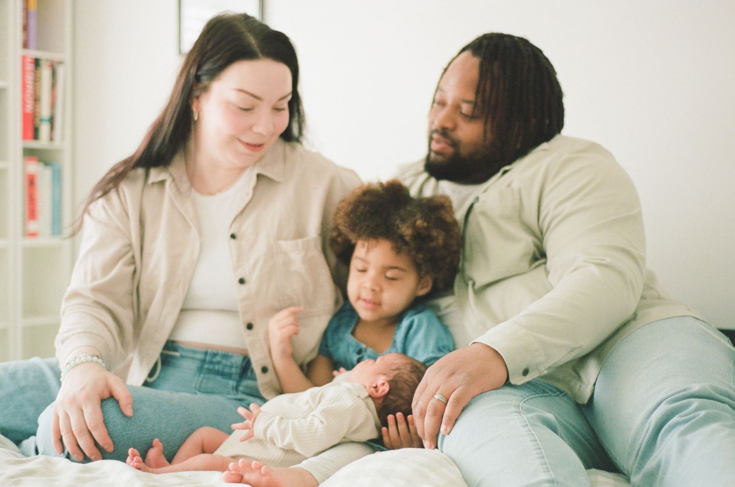 Interracial family with newborn baby and toddler sitting together on a couch in a bright living room.