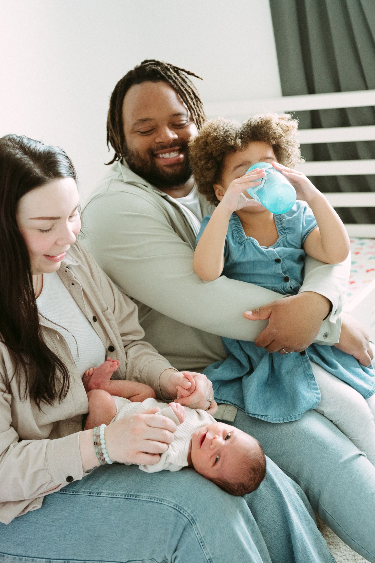Happy interracial family sitting together, parents smiling at newborn baby while toddler drinks from sippy cup.