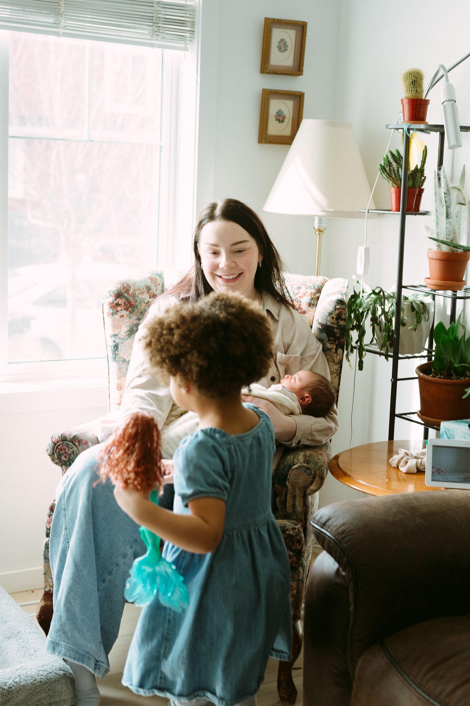 A smiling mother sits in an armchair holding a newborn while a curly-haired toddler in blue dress approaches.