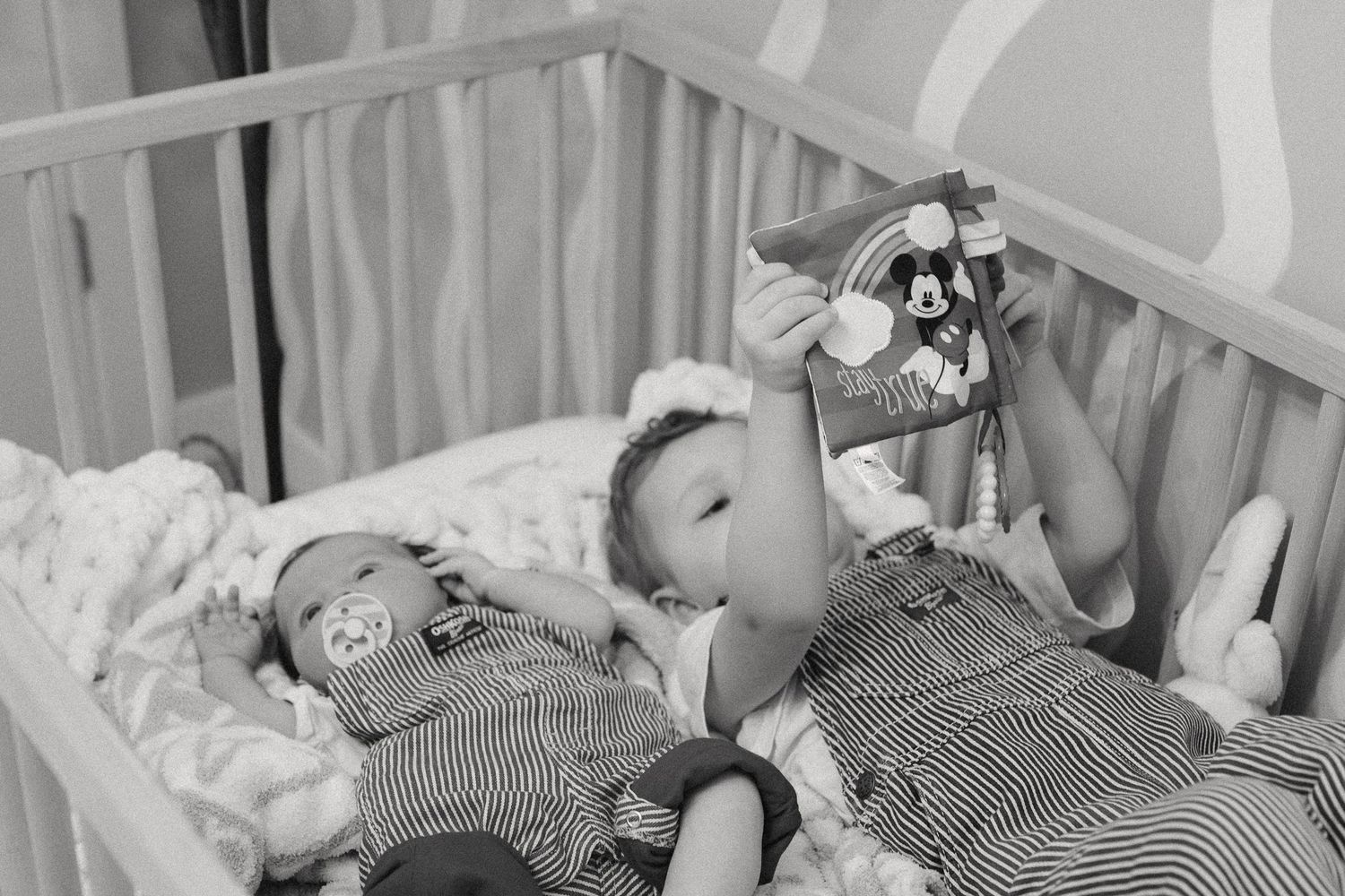 A black and white photo of a baby in striped overalls lying in a crib and playing with a hanging toy.